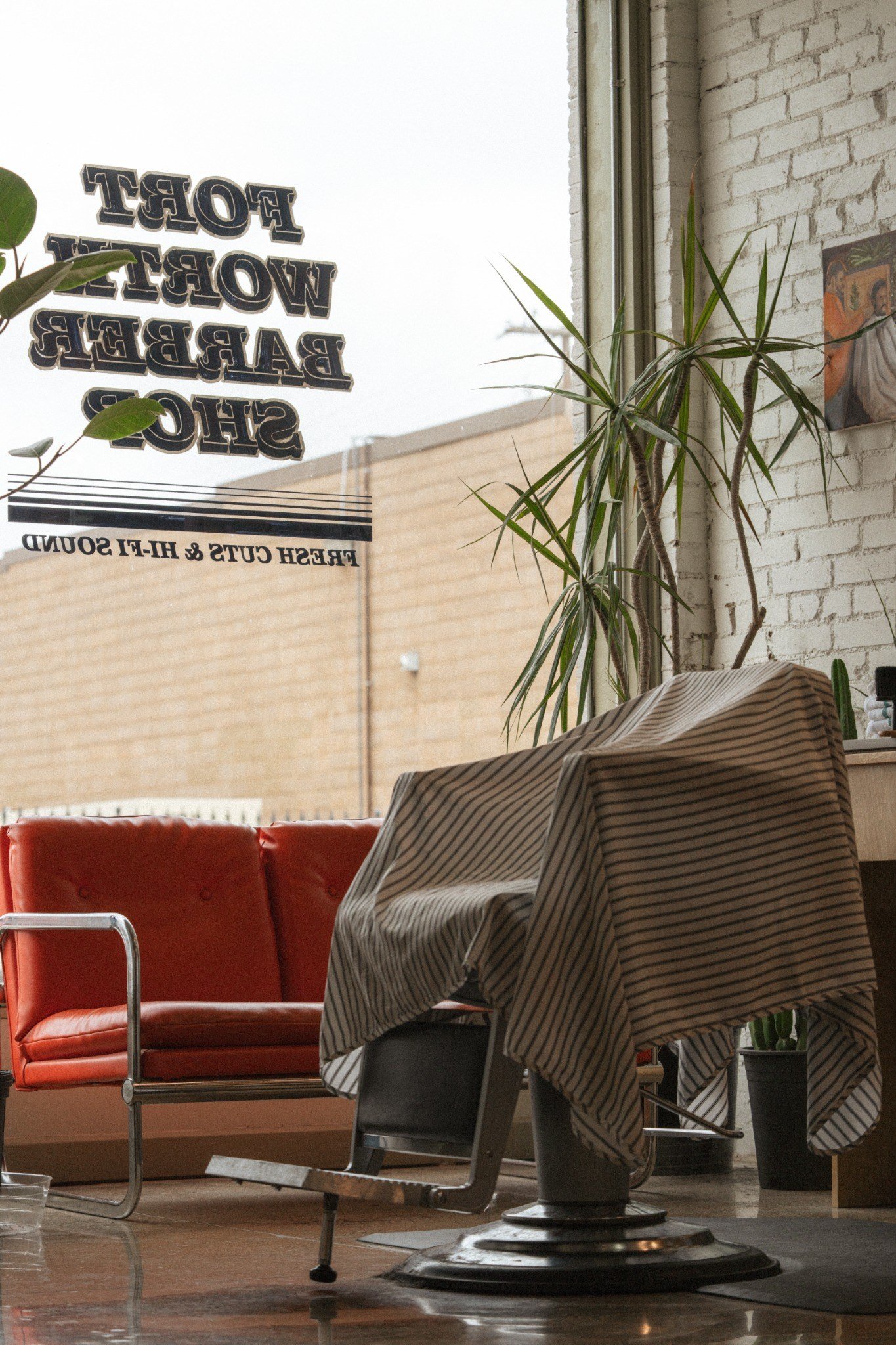 Inside a modern office or cafe with a white brick wall, a large window, plants, a red leather sofa, and a table with a striped tablecloth.
