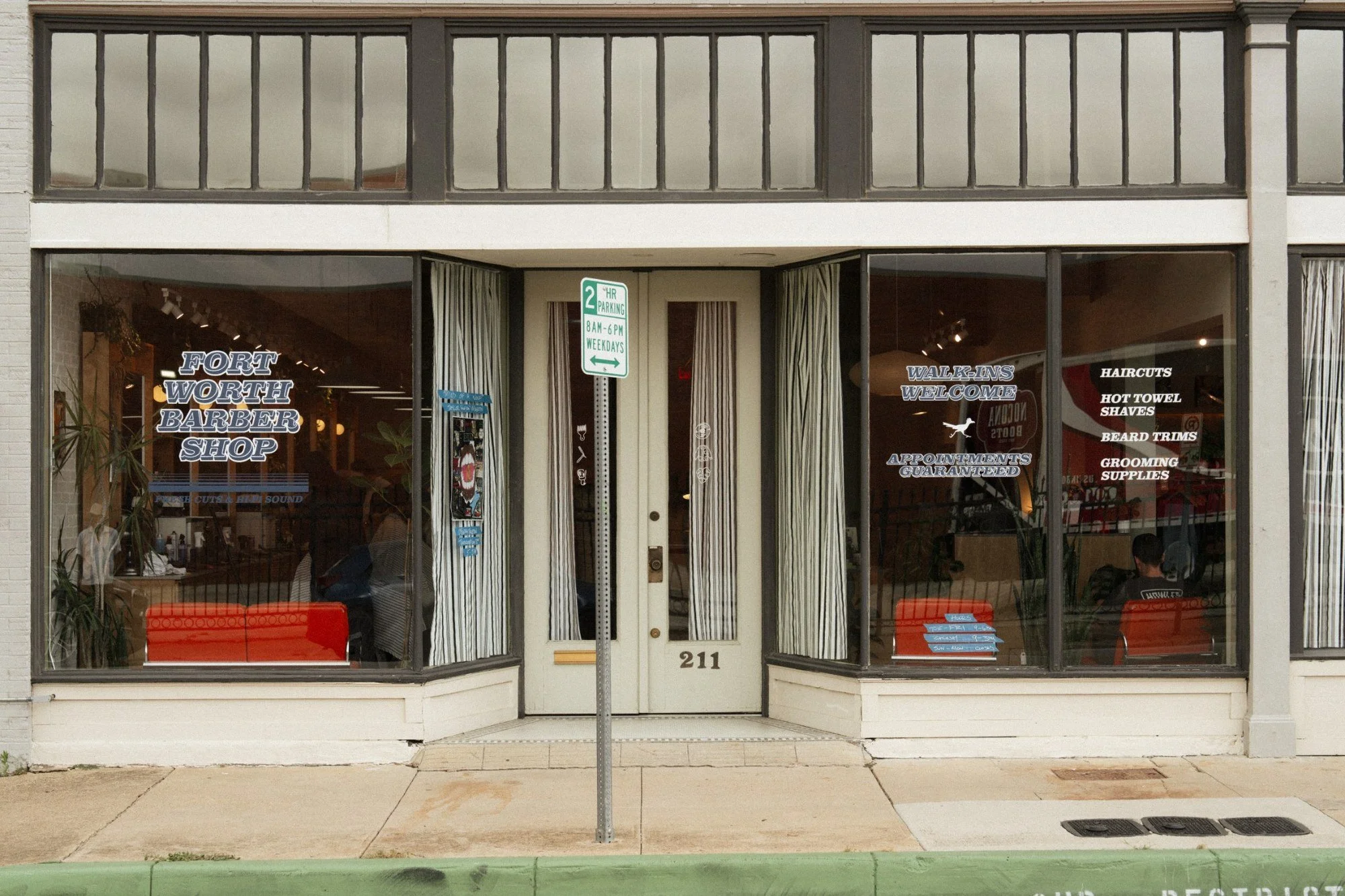 Front view of a barber shop named Fort Worth Barber Shop with large windows displaying signs for walk-ins welcome, appointments guaranteed, and services like haircuts, hot towel shaves, beard trimmings, and grooming supplies. The storefront has a sidewalk in front with a parking sign and some orange benches inside visible through the windows.