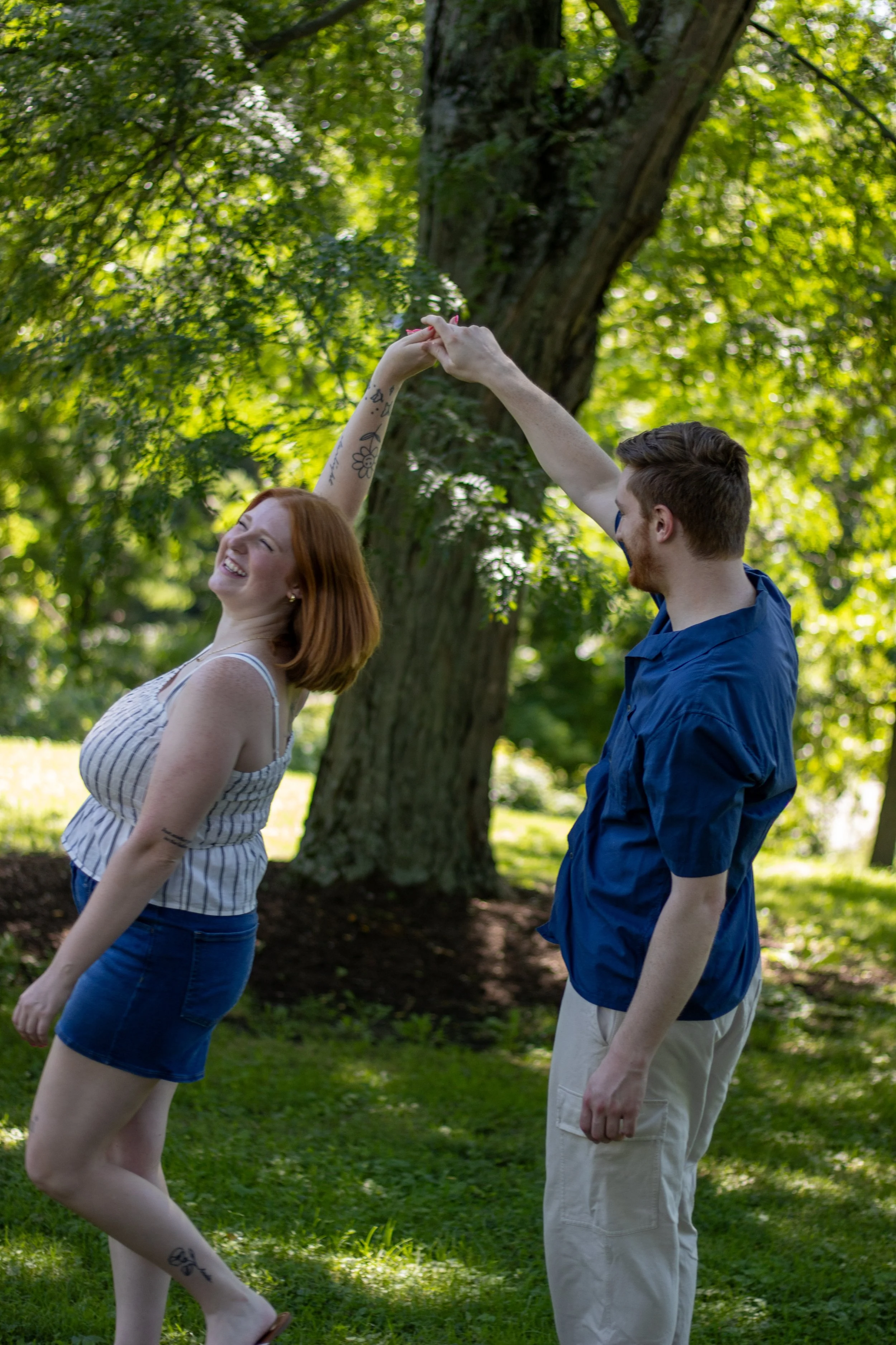 A young couple is dancing outside in a park with green grass and trees. The woman has red hair, tattoos on her arm and leg, and is smiling while looking up. The man has short brown hair, a beard, and is wearing a blue shirt and beige pants. They are 