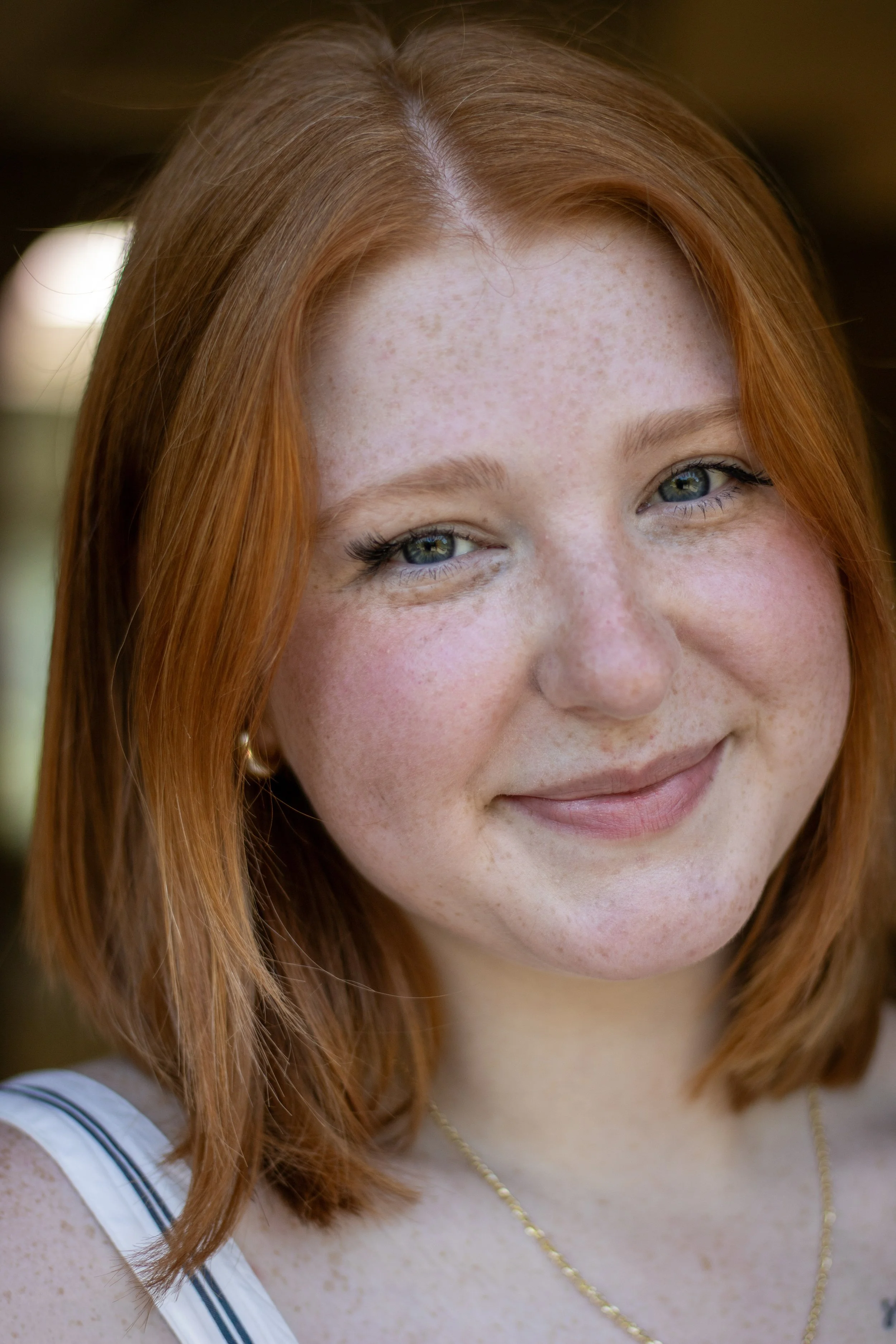 Close-up of a young woman with red hair, green eyes, freckles, and a smiling expression, wearing a white top with black stripes and a gold chain necklace.
