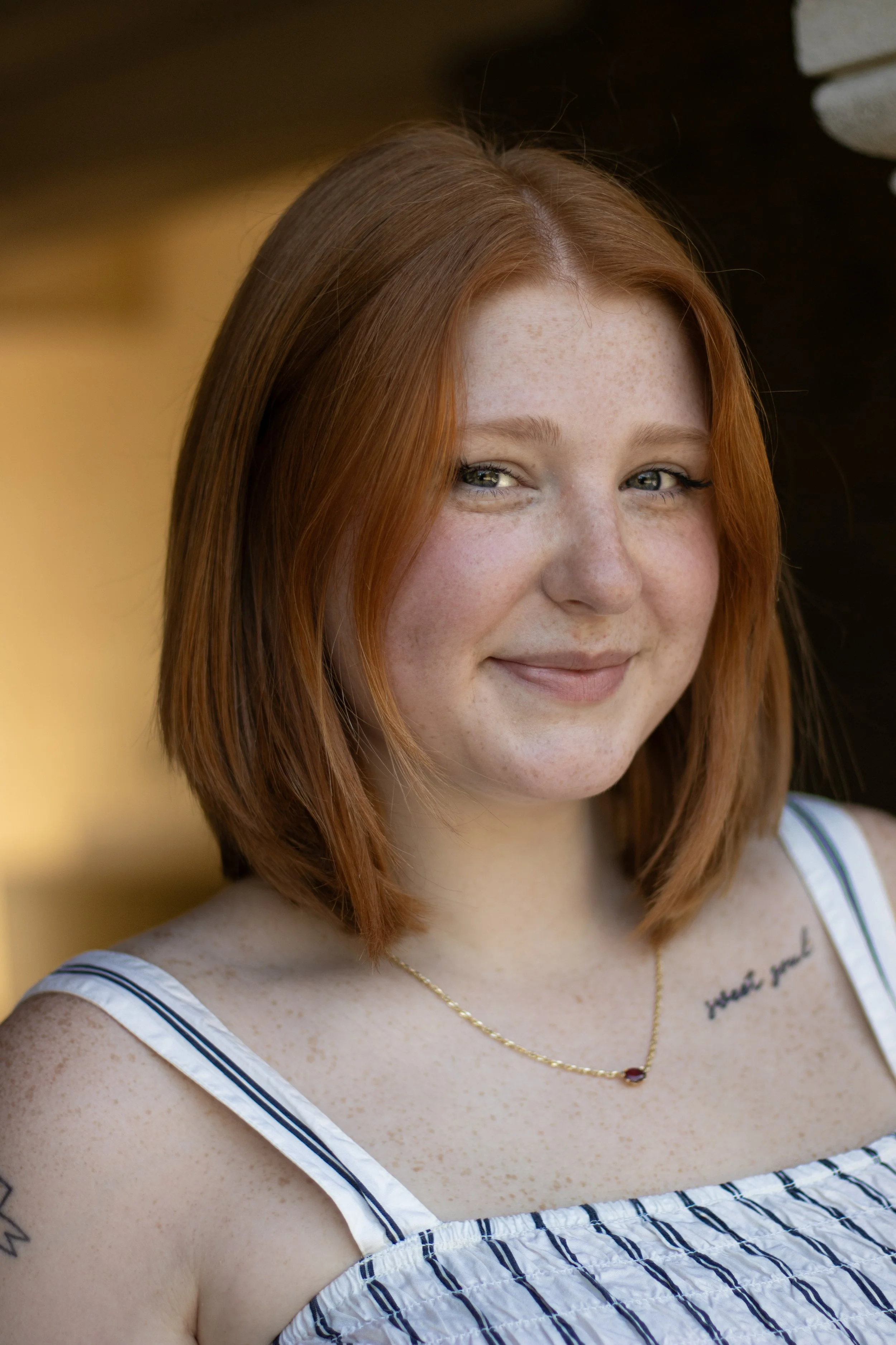 A young woman with red hair, freckles, and blue eyes smiling in front of a blurred yellow and dark background, wearing a striped summer dress and a gold necklace.