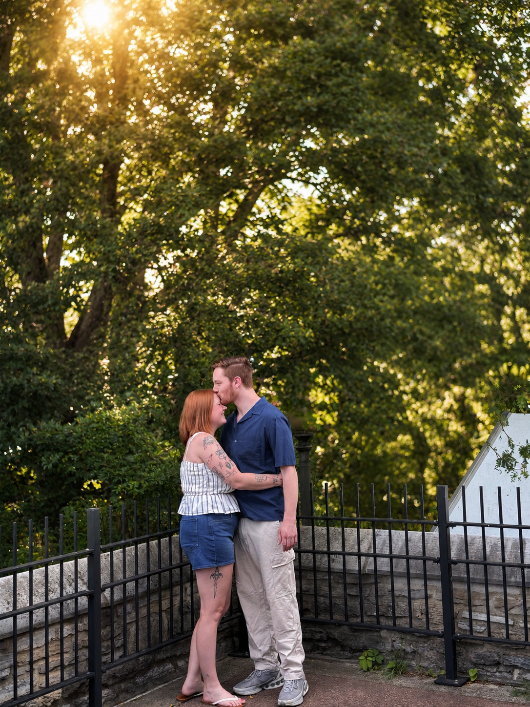 A couple sharing a kiss in a park during sunset, surrounded by trees and a black metal fence.