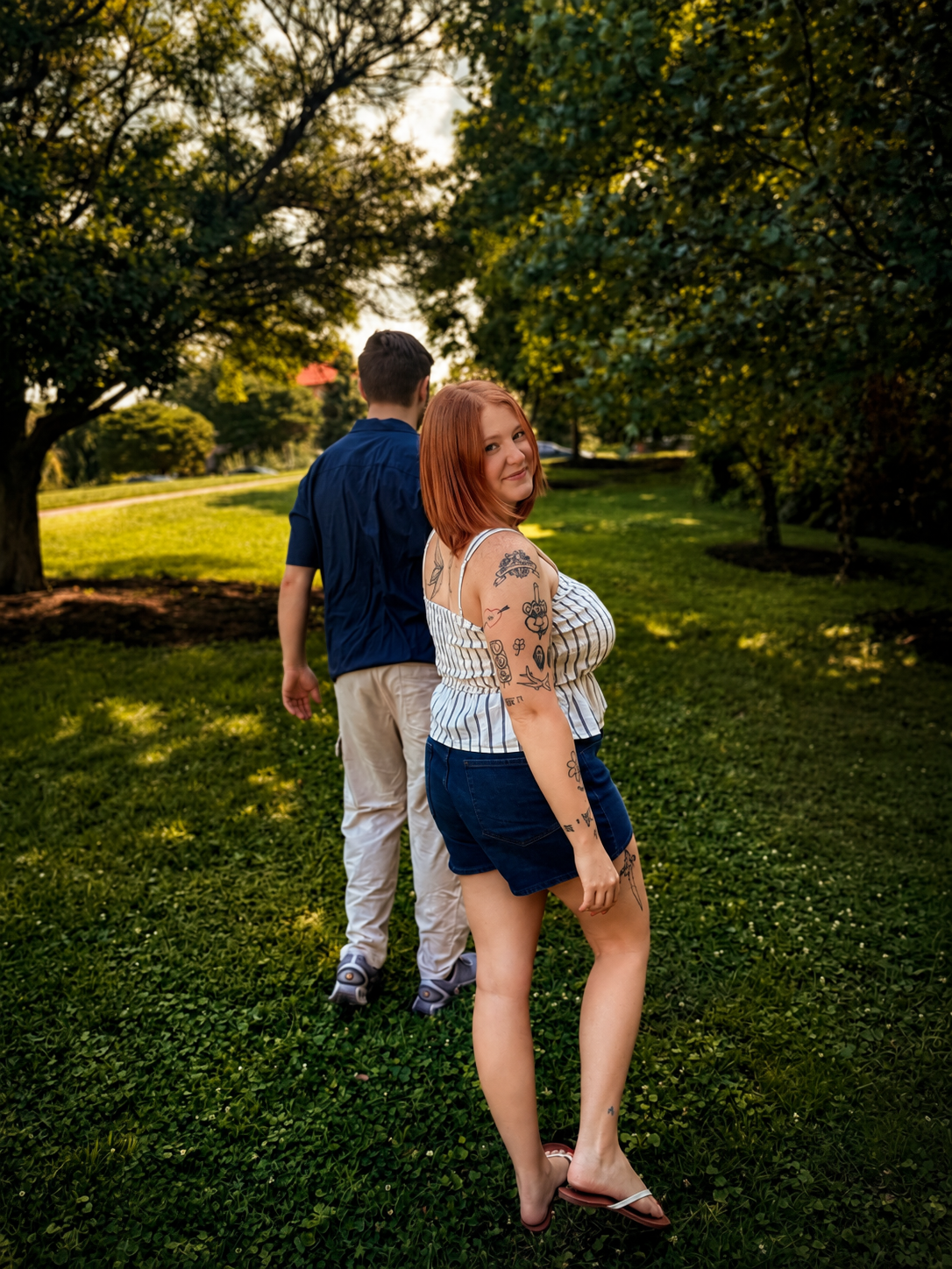 A woman with red hair and tattoos on her arms, wearing a white striped top, denim shorts, and flip-flops, standing on grass in a park, smiling at the camera. A man with brown hair, wearing a dark blue shirt and light-colored pants, is walking ahead w