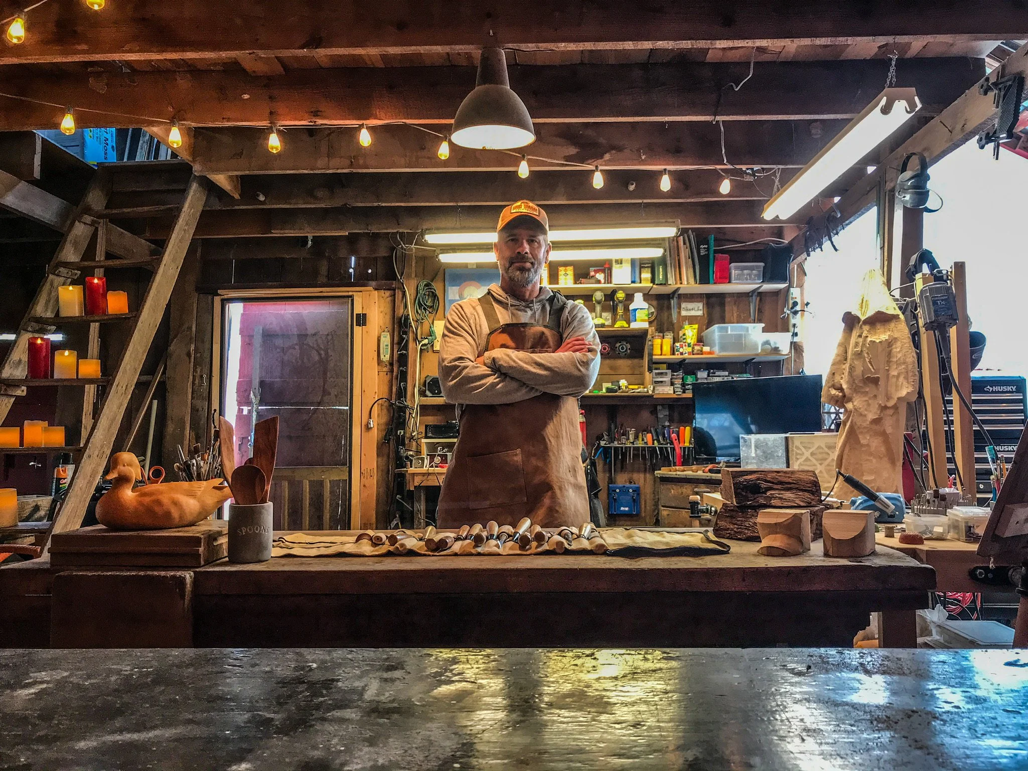 A man stands behind a woodworking workbench in a rustic workshop, surrounded by tools, wood pieces, and decorative candles.