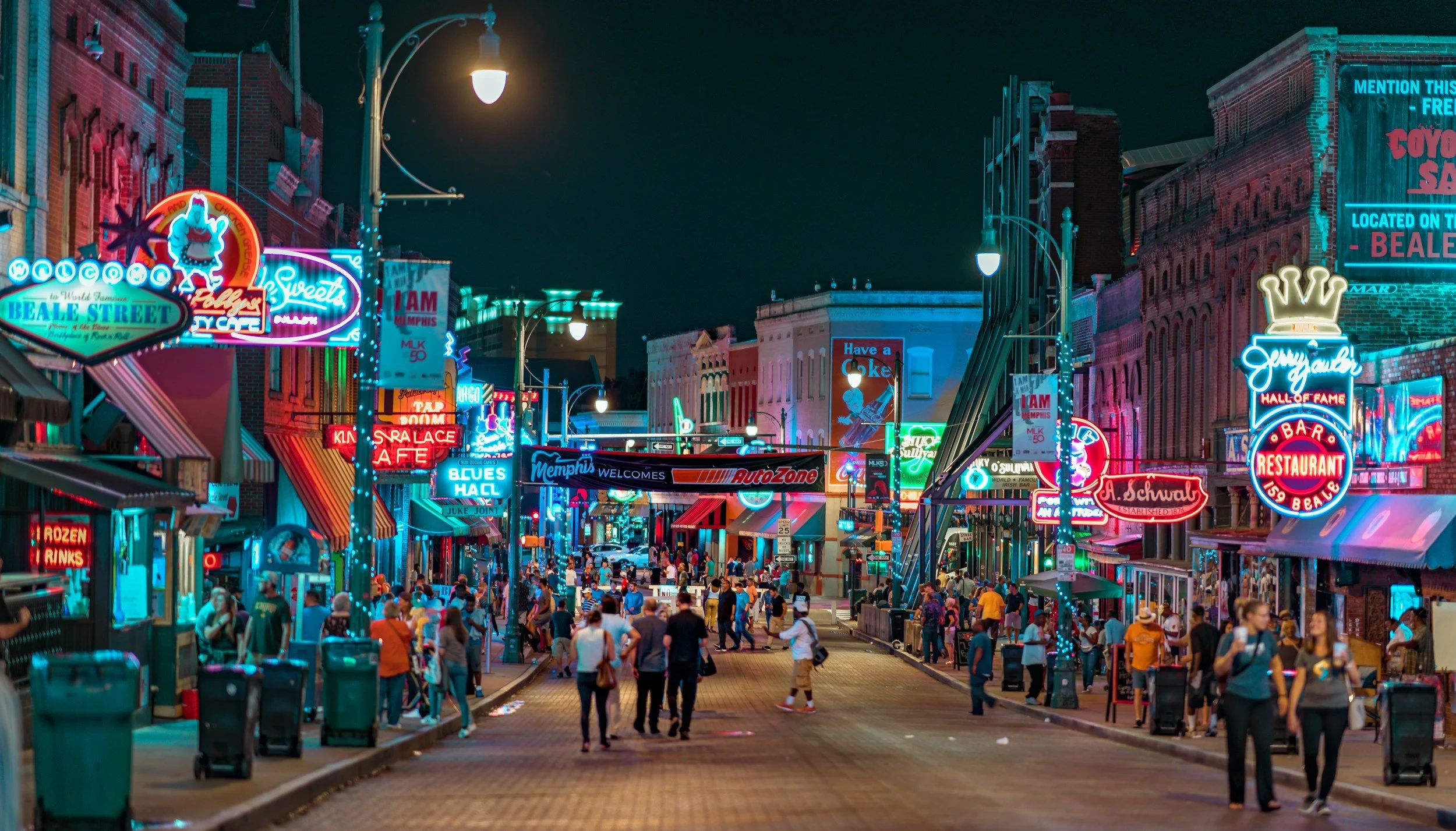 Nighttime street scene on Beale Street in Memphis, decorated with colorful neon signs for bars, restaurants, and entertainment venues, with a crowd of people walking and socializing.