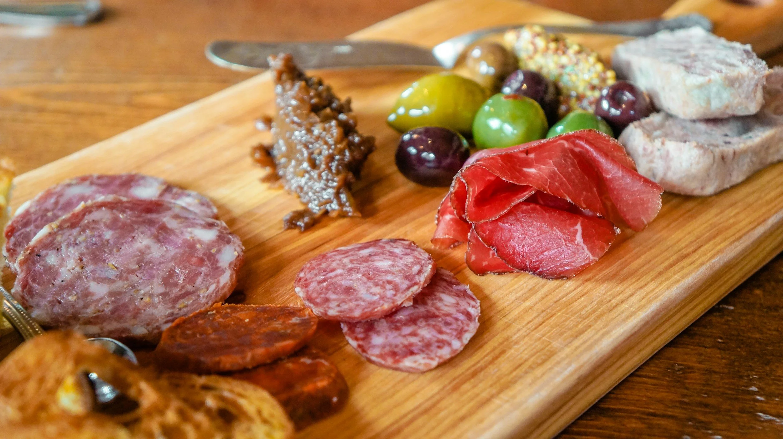 Assorted slices of cured meats and a selection of green and purple olives on a wooden serving board.