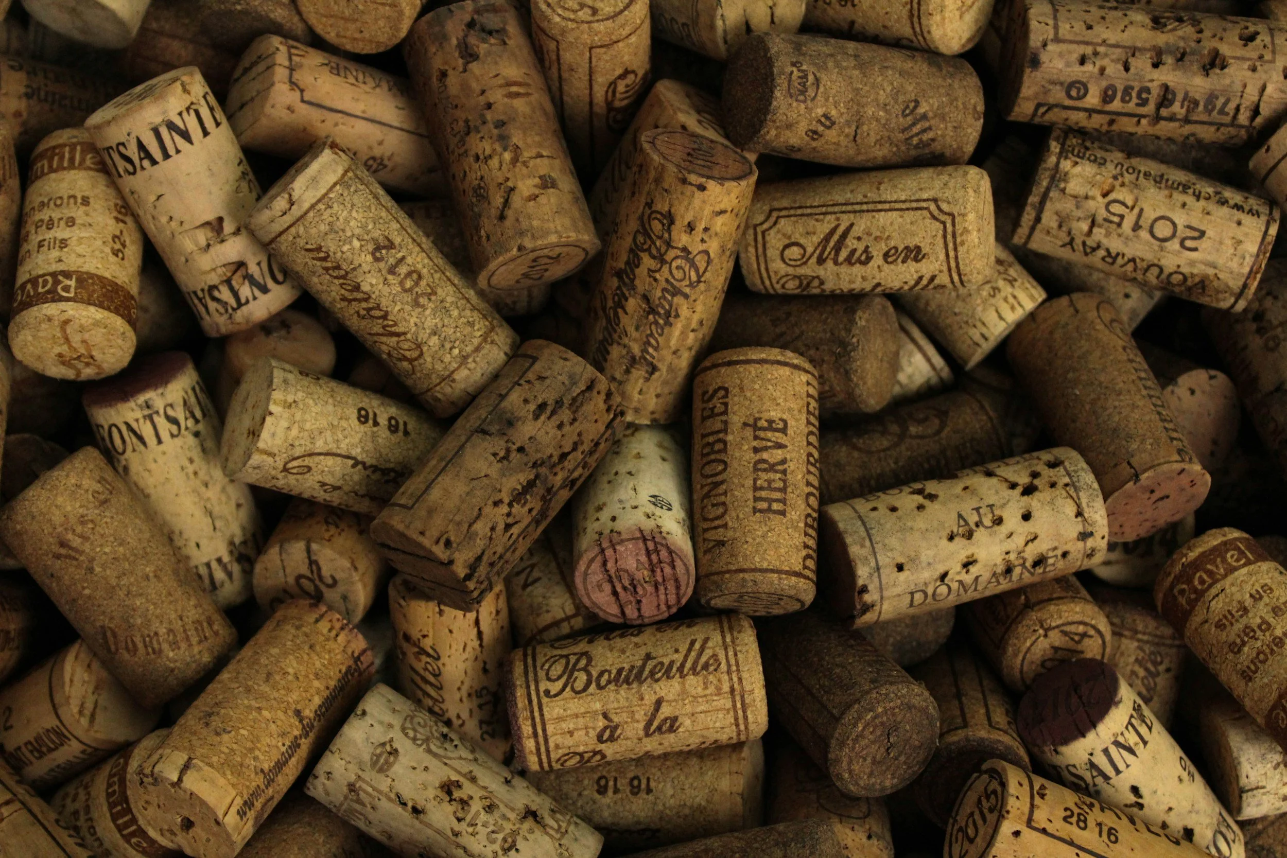 Close-up of numerous used wine corks, some with visible text and markings. The corks are scattered and show signs of wear, with some ink markings and stamps.