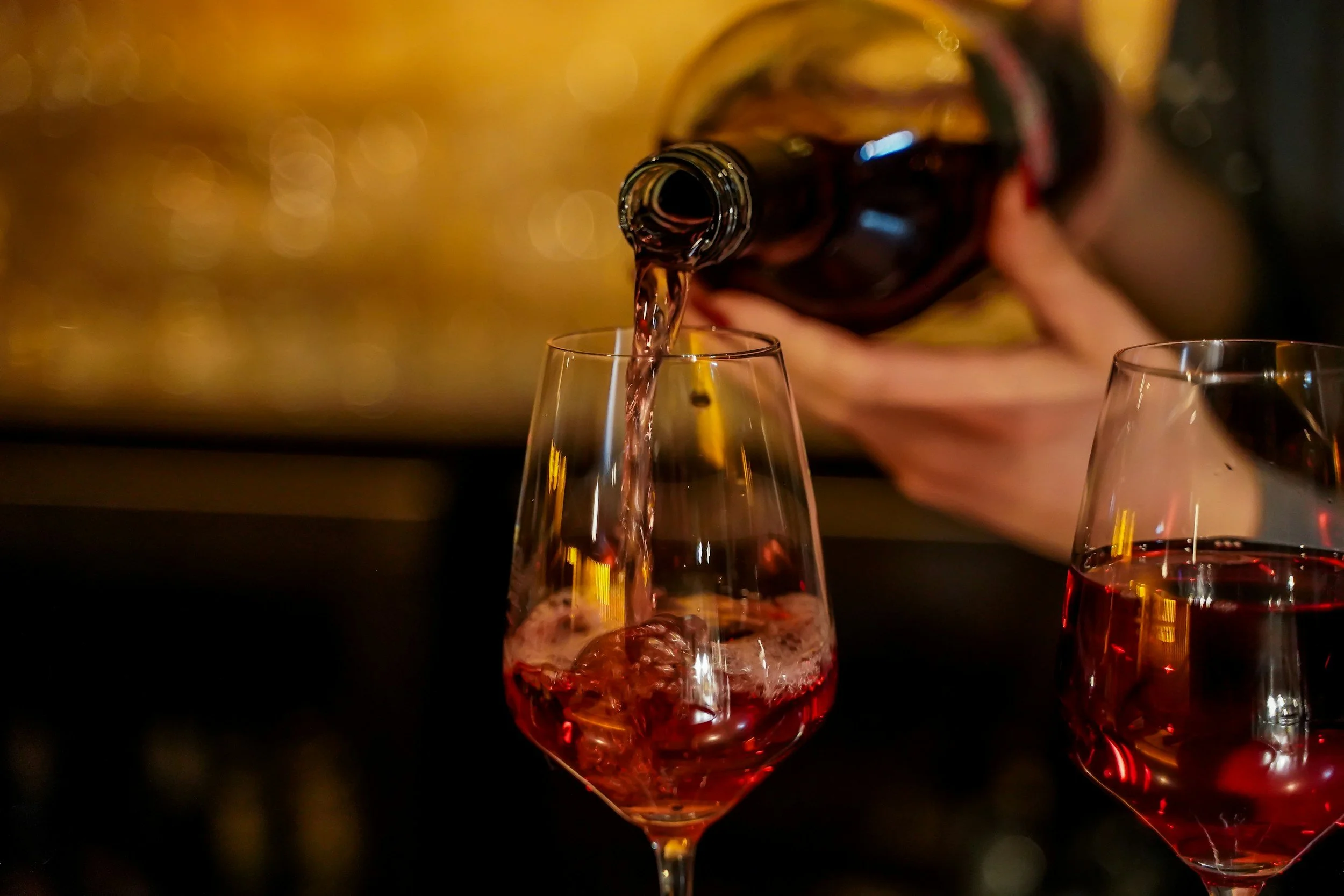 A person pouring red wine from a bottle into a wine glass, with another glass of red wine visible in the foreground, in a warm, bokeh background.