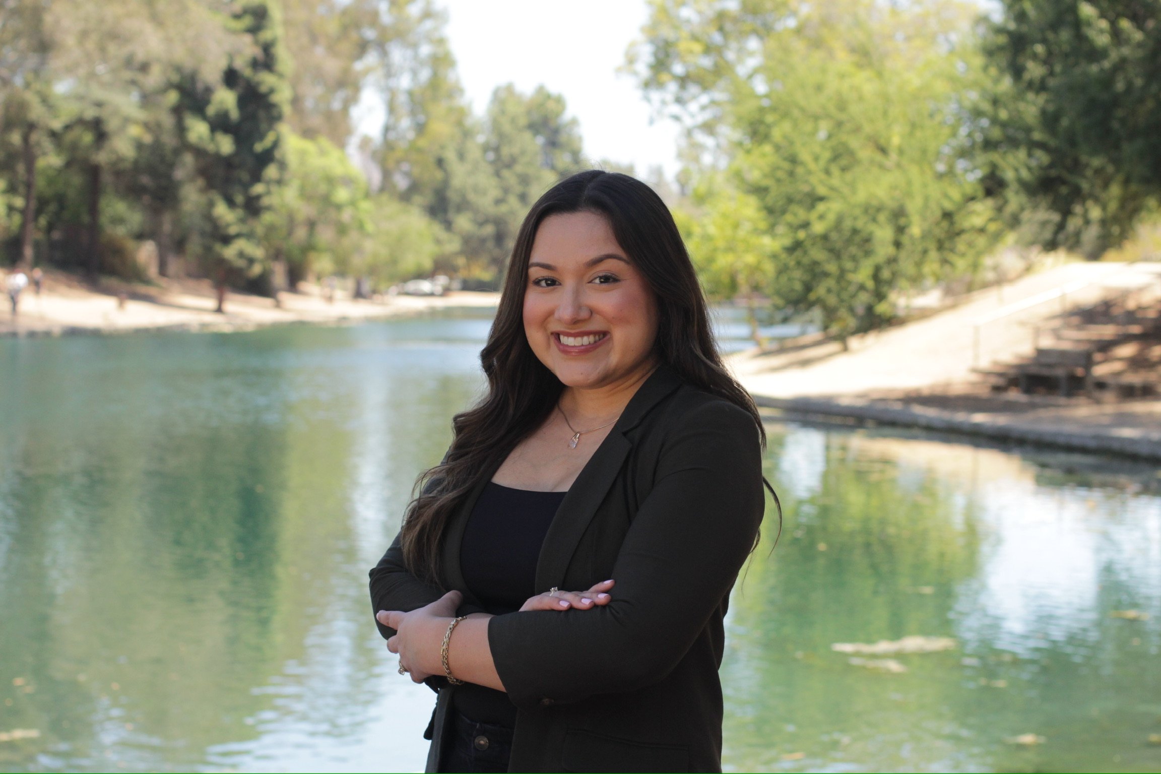 A woman with long dark hair, wearing a black blazer, standing near a body of water with trees and a park background.