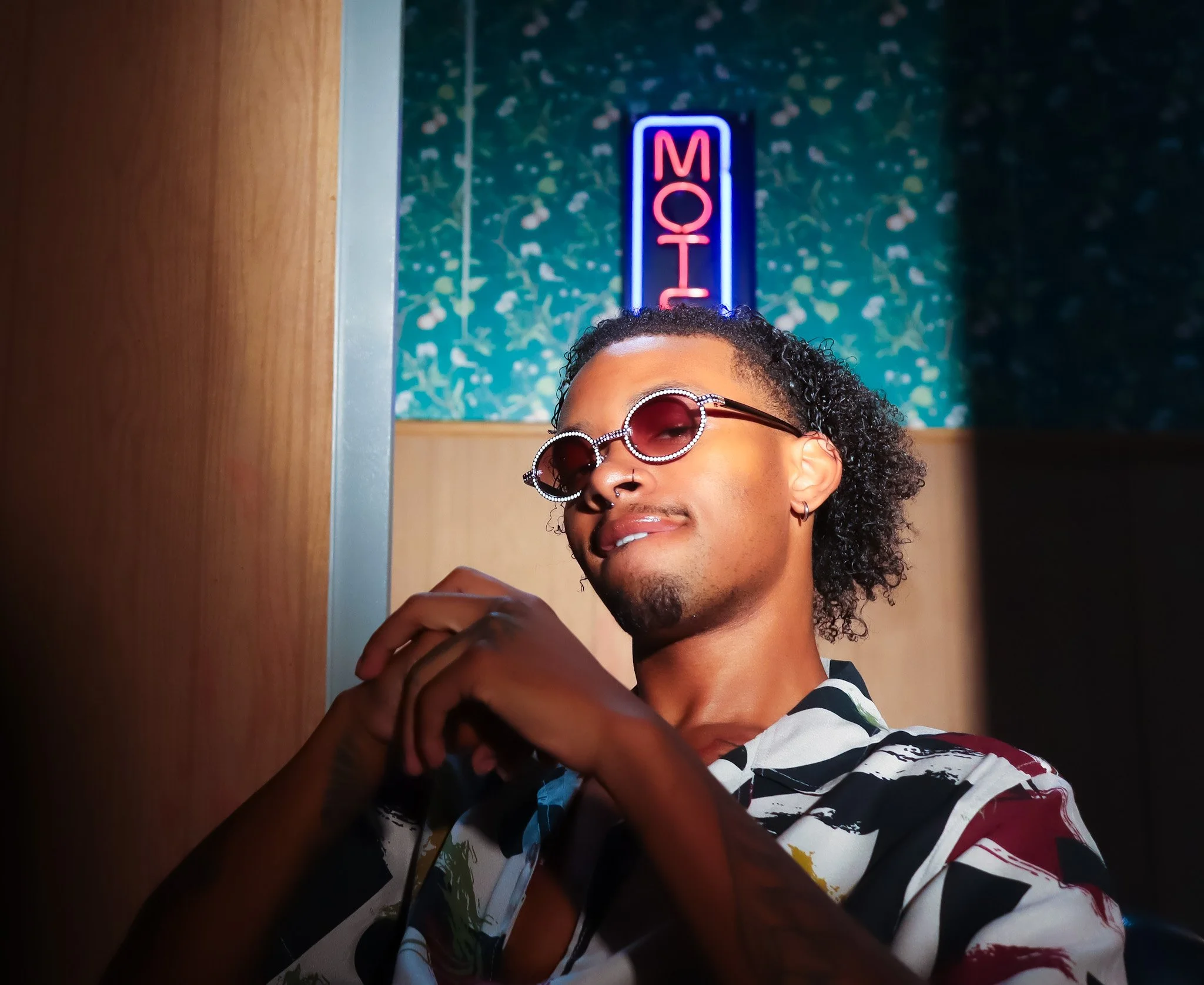 A young man with curly hair wearing sunglasses and a colorful patterned shirt, reaching out and smiling as he looks down. Behind him, there is a neon sign that says 'MOV' and a background with a green leafy pattern. 90s motel scene
