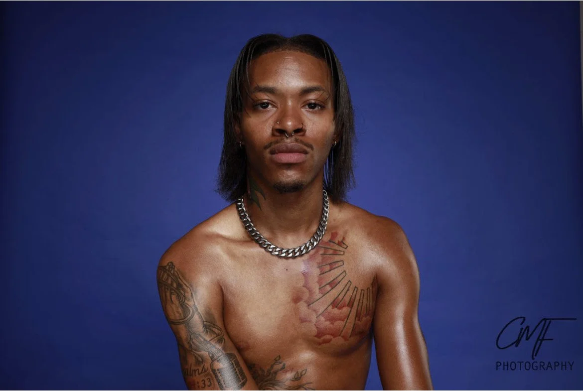 Portrait of a shirtless man with tattoos, a silver chain necklace, and dark hair, against a blue background. Prince inspired photoshoot