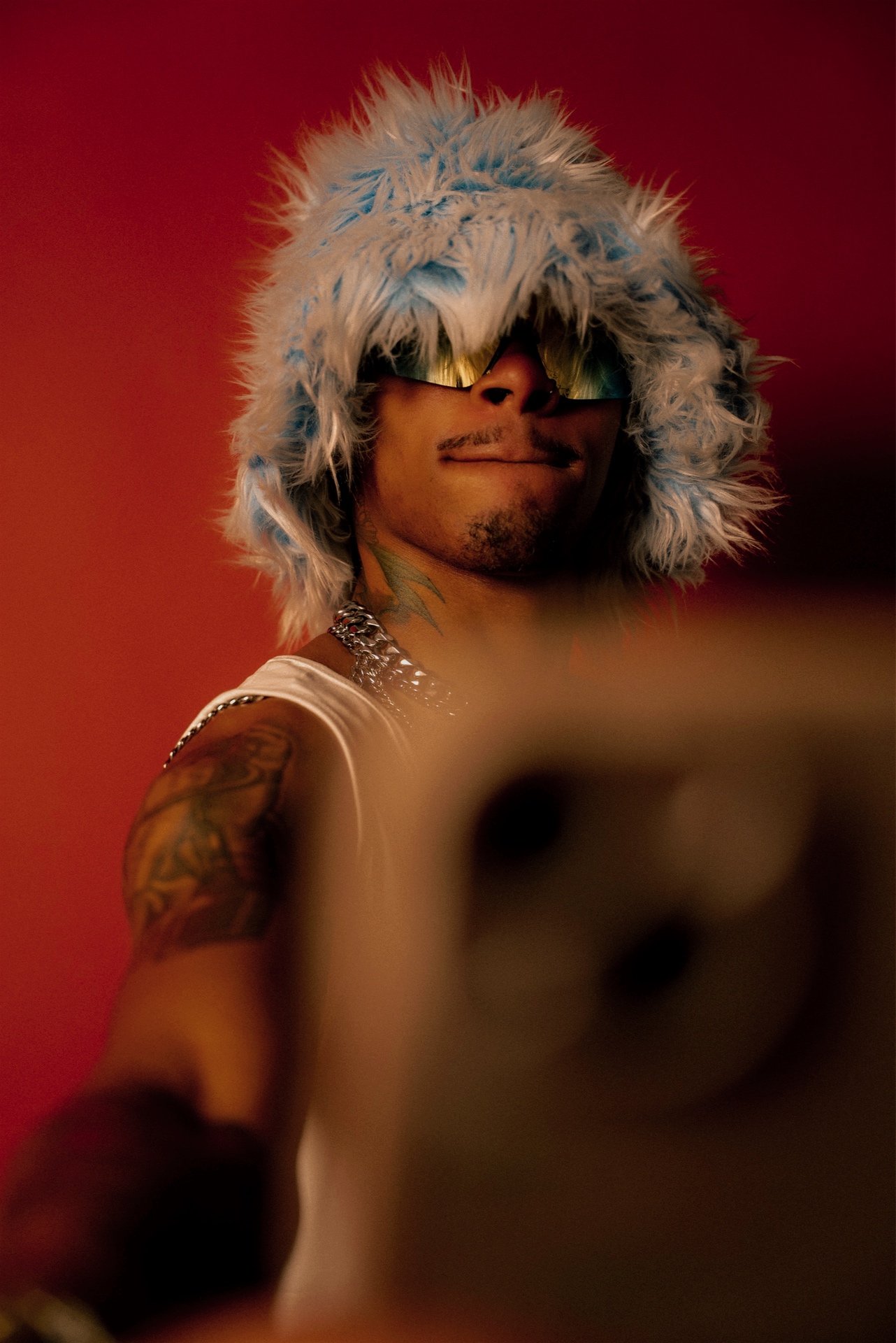 A person wearing a fluffy white and blue hood, sunglasses, and a white tank top, taking a selfie against a red background.