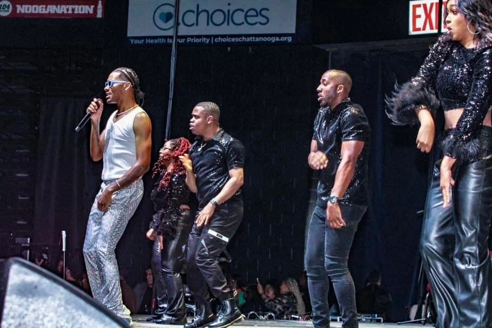 Performers dancing on stage during a concert, with five individuals dressed in stylish outfits, some holding microphones.