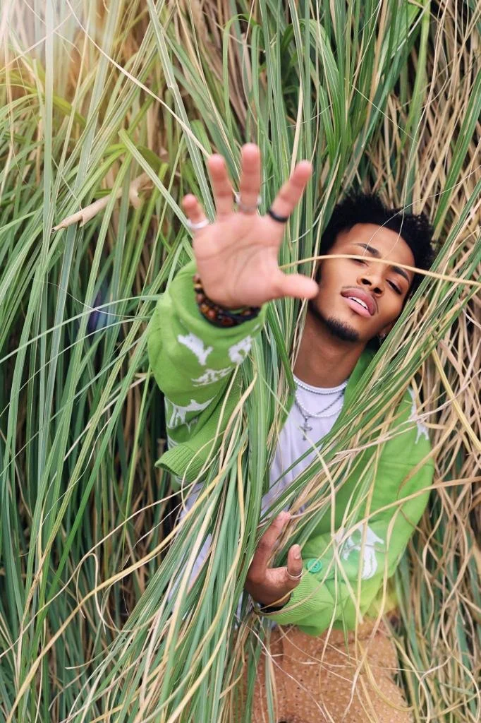 A person with black curly hair and jewelry, wearing a green sweater and white shirt, is reaching out through tall grass, with their face partly hidden among the grass. Nature photoshoot, immersive inspiration