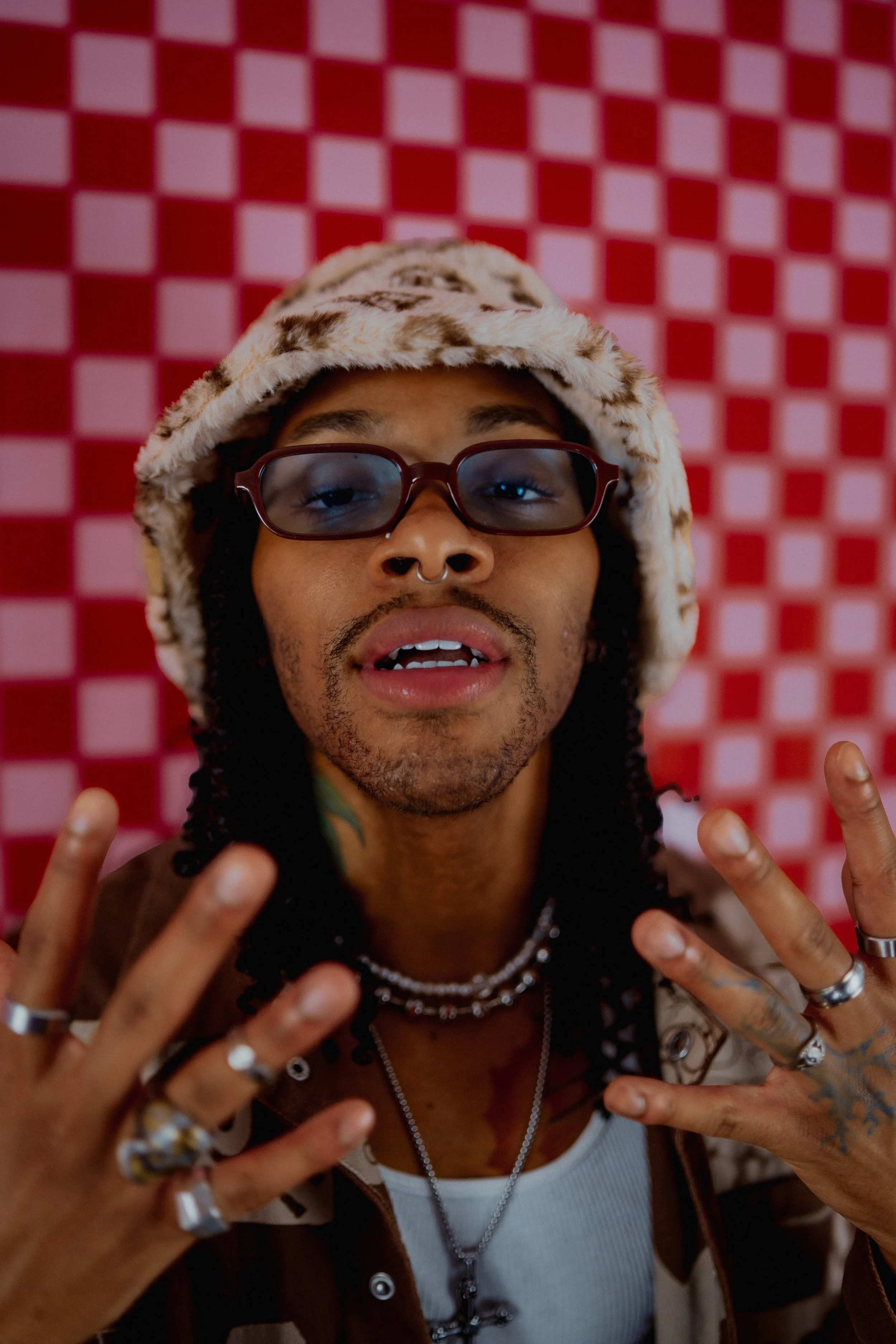 A person with dreadlocks, wearing glasses, a fur hat, multiple rings, necklaces, and a septum piercing is making a peace sign with both hands in front of a red and white checkered wall. Creative inspiration for models