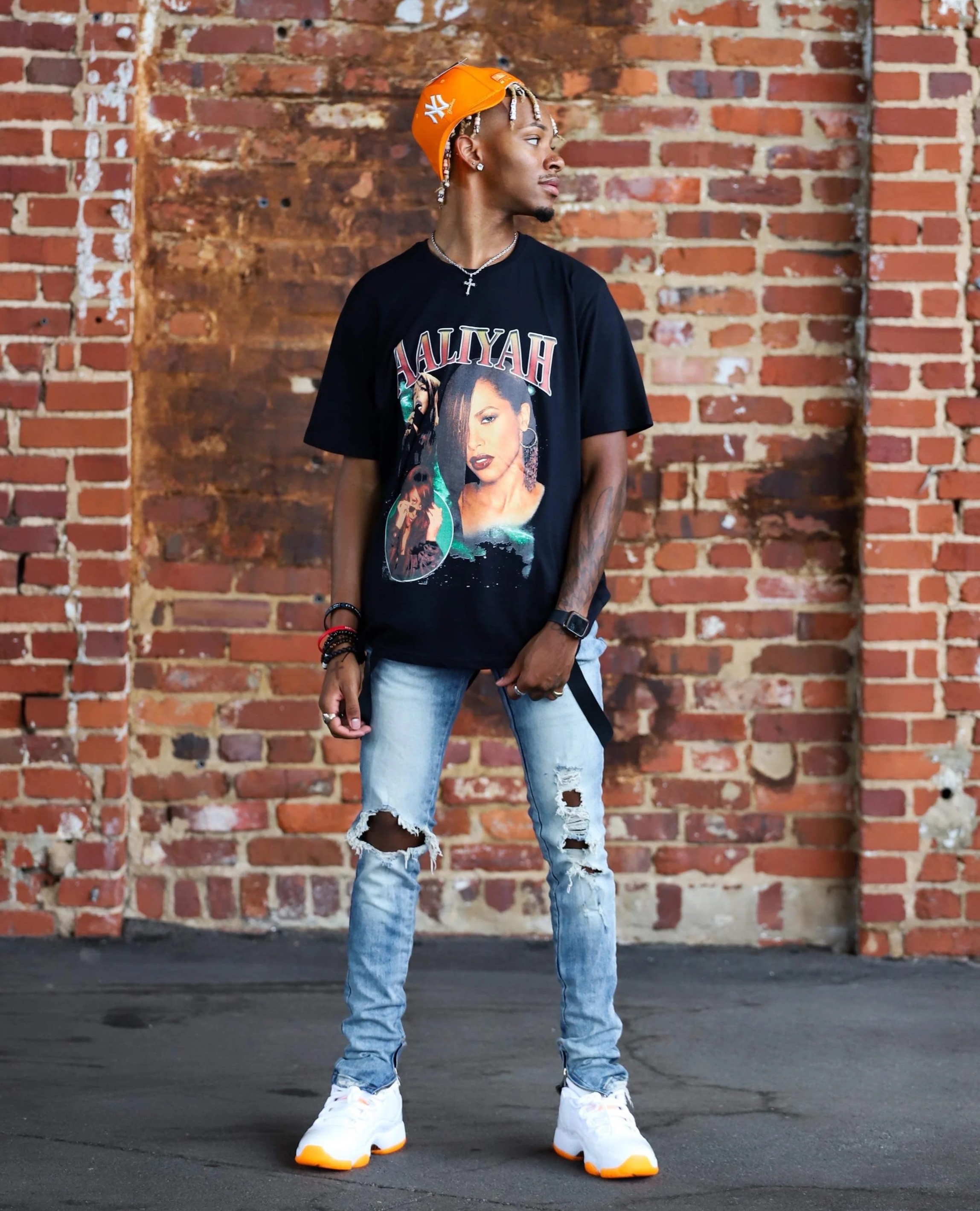 A young man wearing a black graphic T-shirt, distressed jeans, white sneakers, and an orange cap backward, stands in front of a red brick wall, looking to the right.
