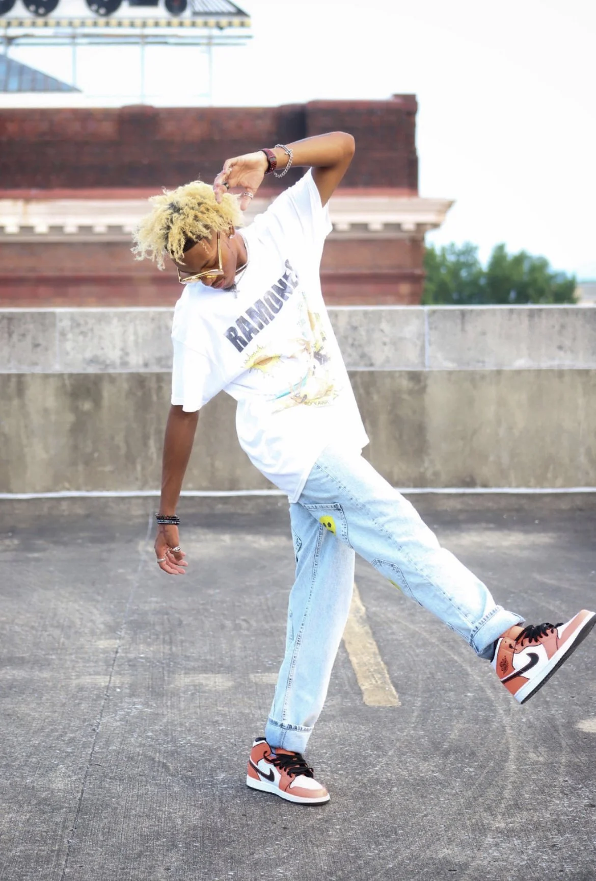 A person with blonde, curly hair wearing glasses, a white Ramones T-shirt, light blue jeans, and Air Jordan sneakers, dancing on a rooftop parking lot with a brick building and trees in the background. Urban fashion and modeling inspiration