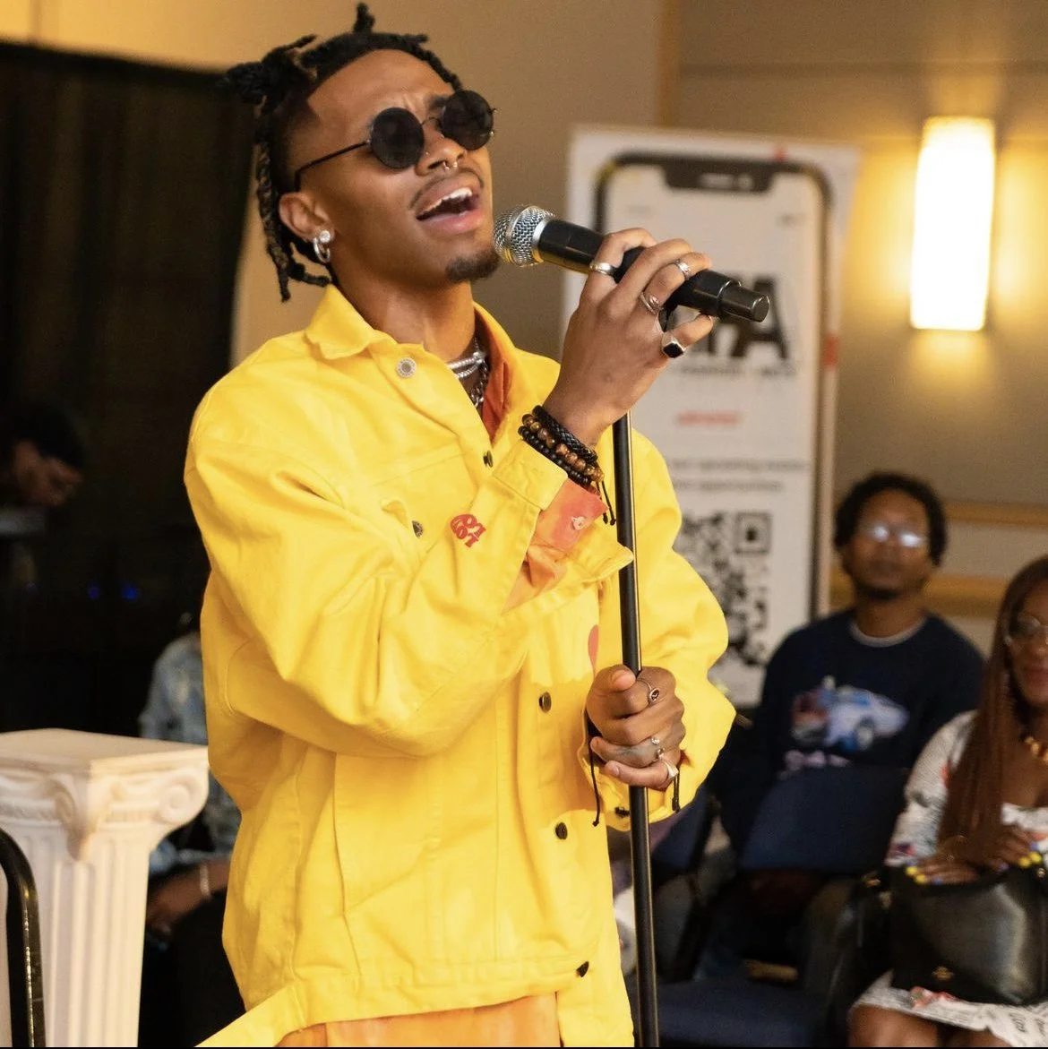 A man singing into a microphone at an indoor event, wearing a bright yellow jacket, black sunglasses, and jewelry. People are seated in the background, and there is a QR code on a poster behind him. Live music, fashion inspiration