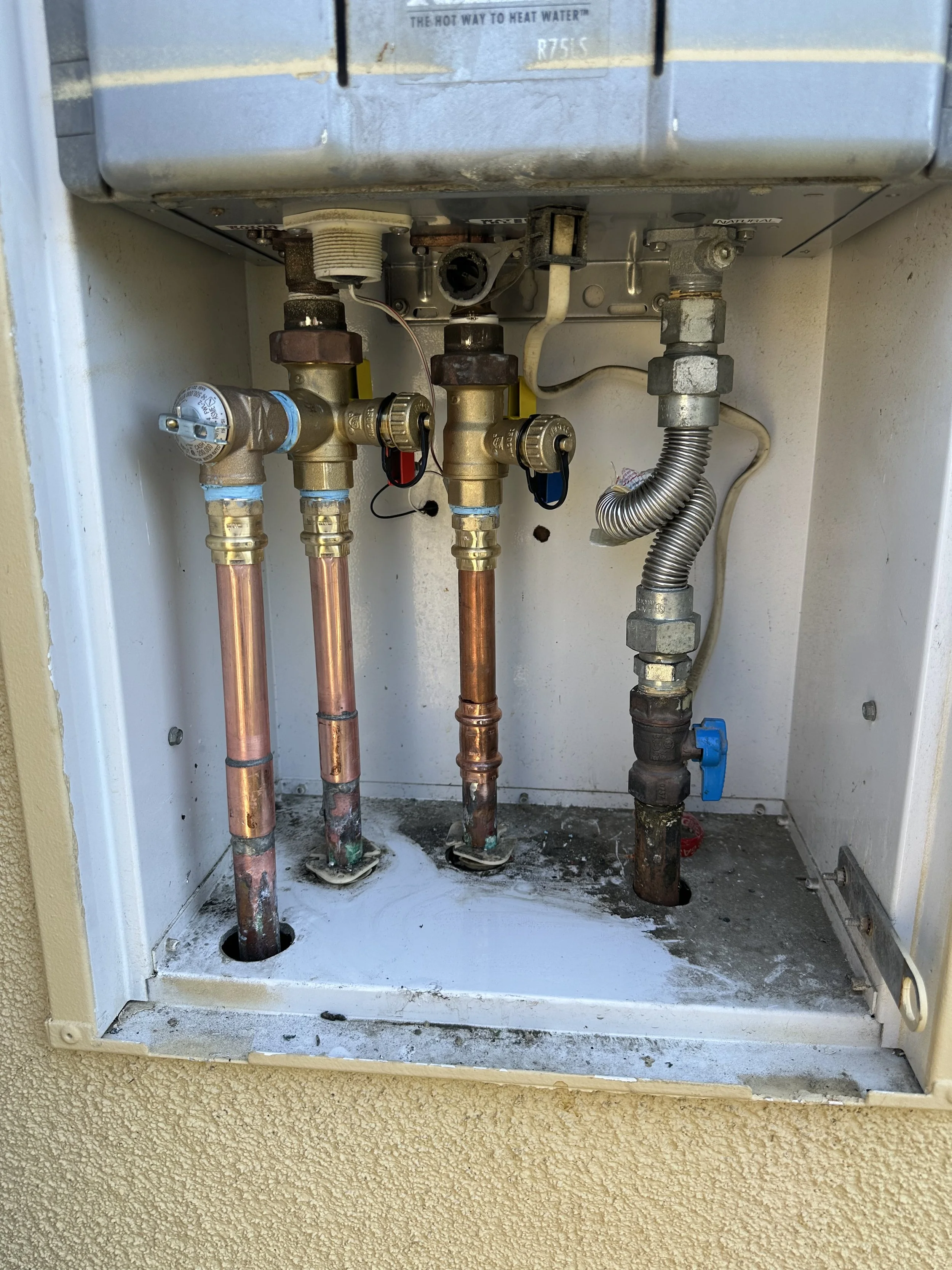 View inside an outdoor electrical box showing plumbing pipes, valves, and wiring, with some corrosion and dirt visible.