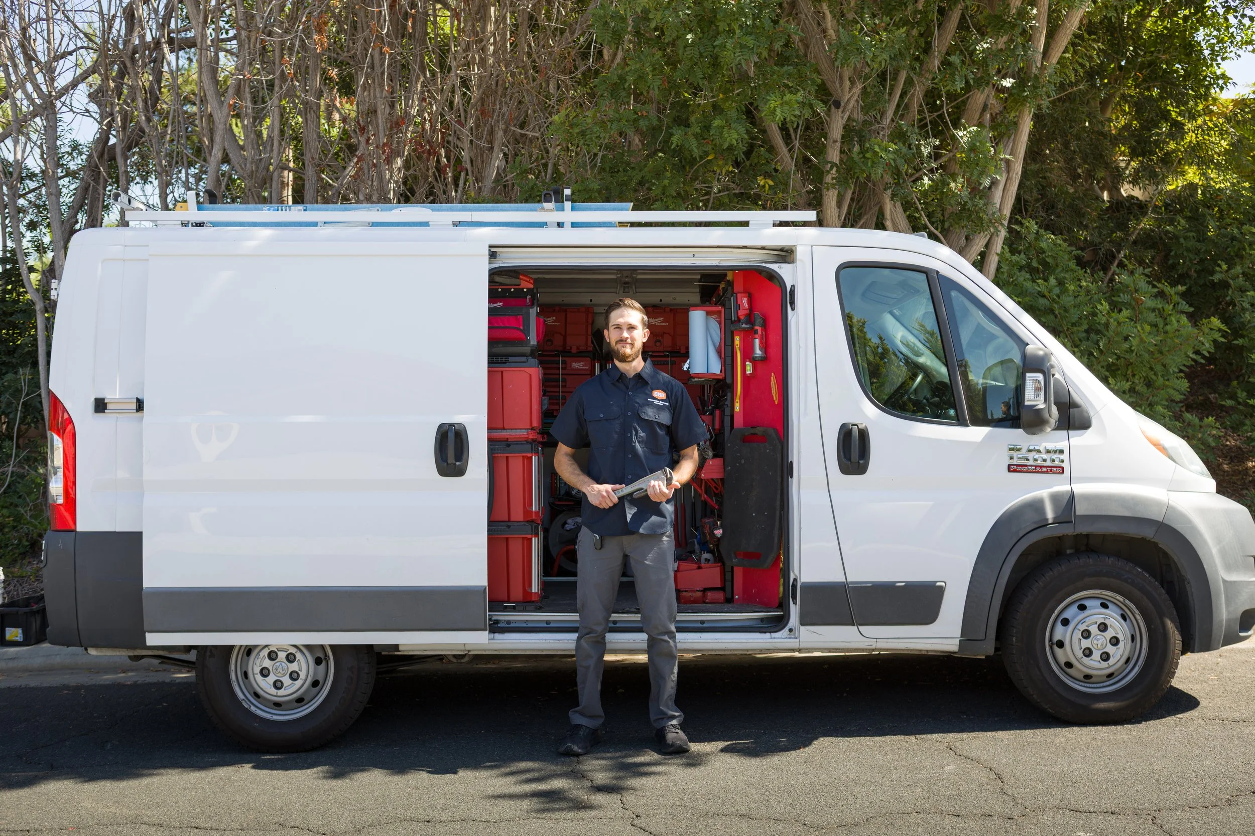 A man with a beard standing in front of a white service van with an open sliding door, revealing tools and equipment inside, parked on a street with trees in the background.