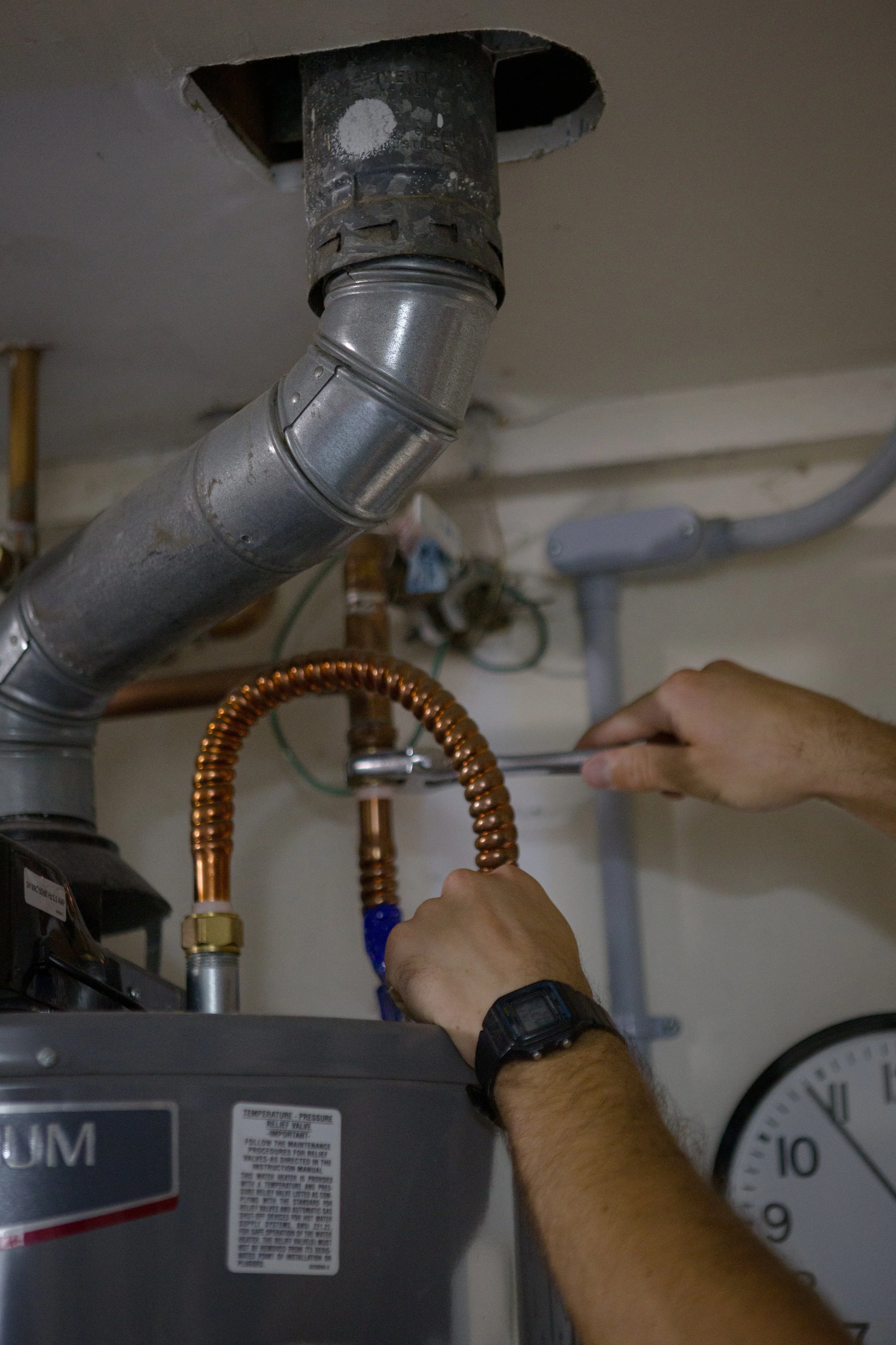 Person using a wrench to service a water heater with pipes and a pressure gauge nearby.