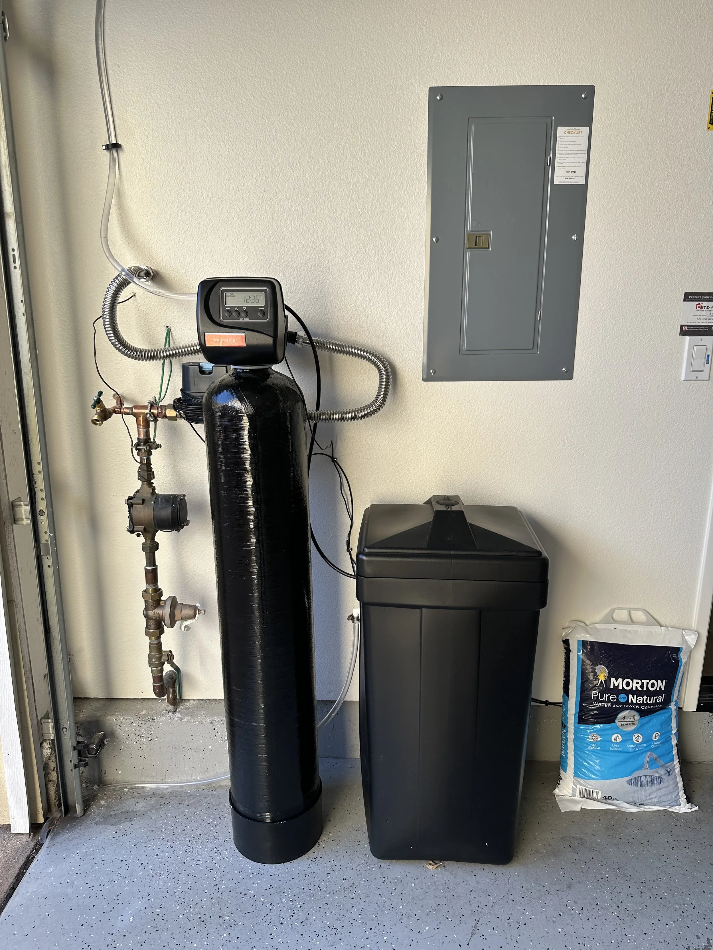 A utility area showing a water softening system, including a black tank with a digital control head, an inlet and outlet pipe, a gray electrical panel on the wall, a black water softener unit, a bag of Morton water softener crystals, and a small trash can on a speckled gray floor.