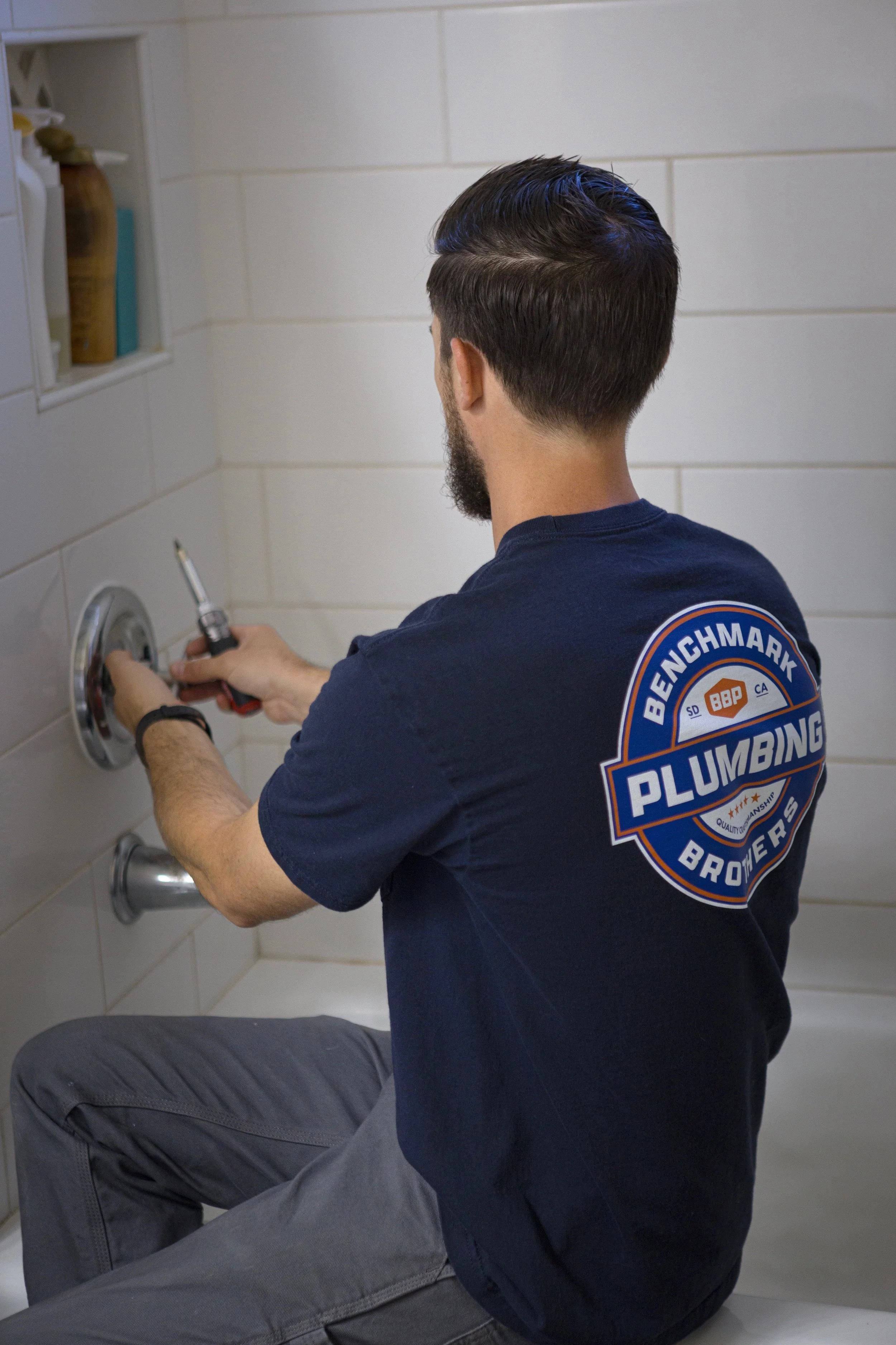 A technician with a Benckmark Plumbing patch on his shirt is kneeling on the floor and working on a plumbing fixture in a tiled bathroom.