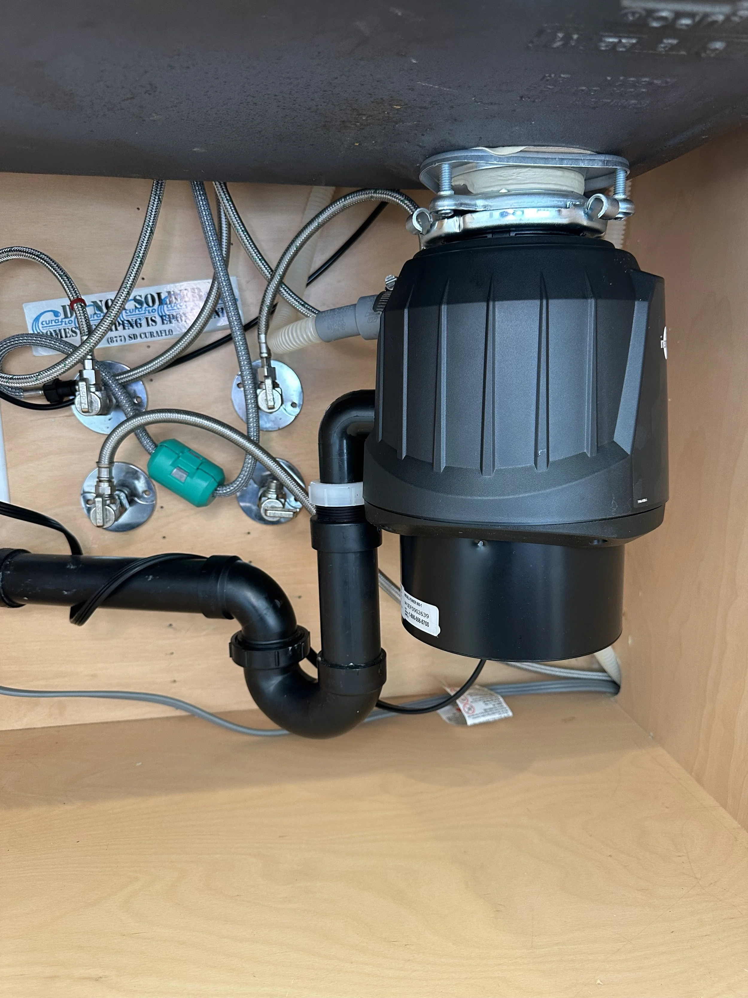 Underneath kitchen sink with plumbing, hoses, and a black garbage disposal unit.