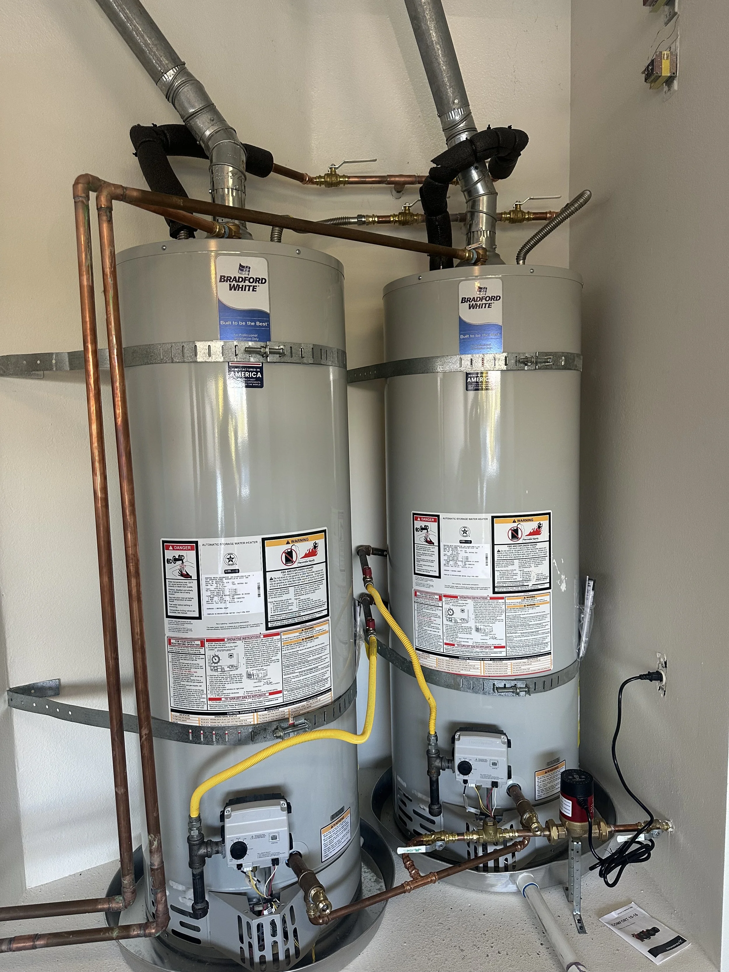 Two large Bradford White water heater tanks connected by copper and flexible piping, installed in a utility room with wiring and a control panel mounted on the wall.