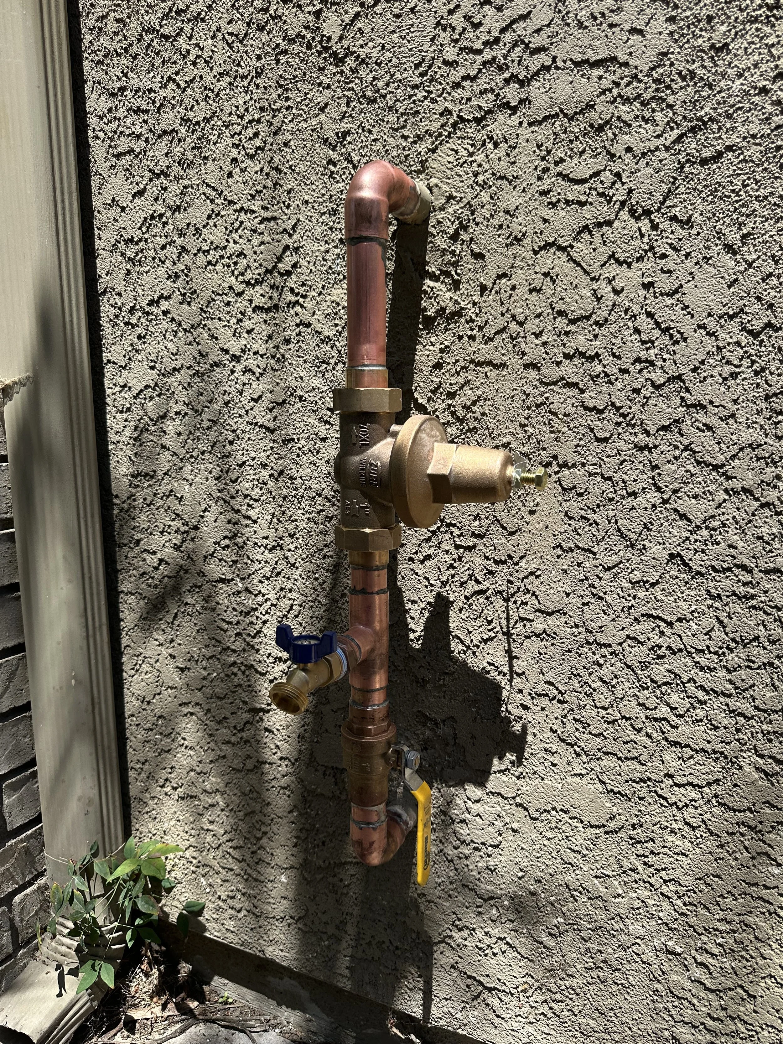Copper plumbing pipe with shut-off valve and fittings mounted on a textured exterior wall.