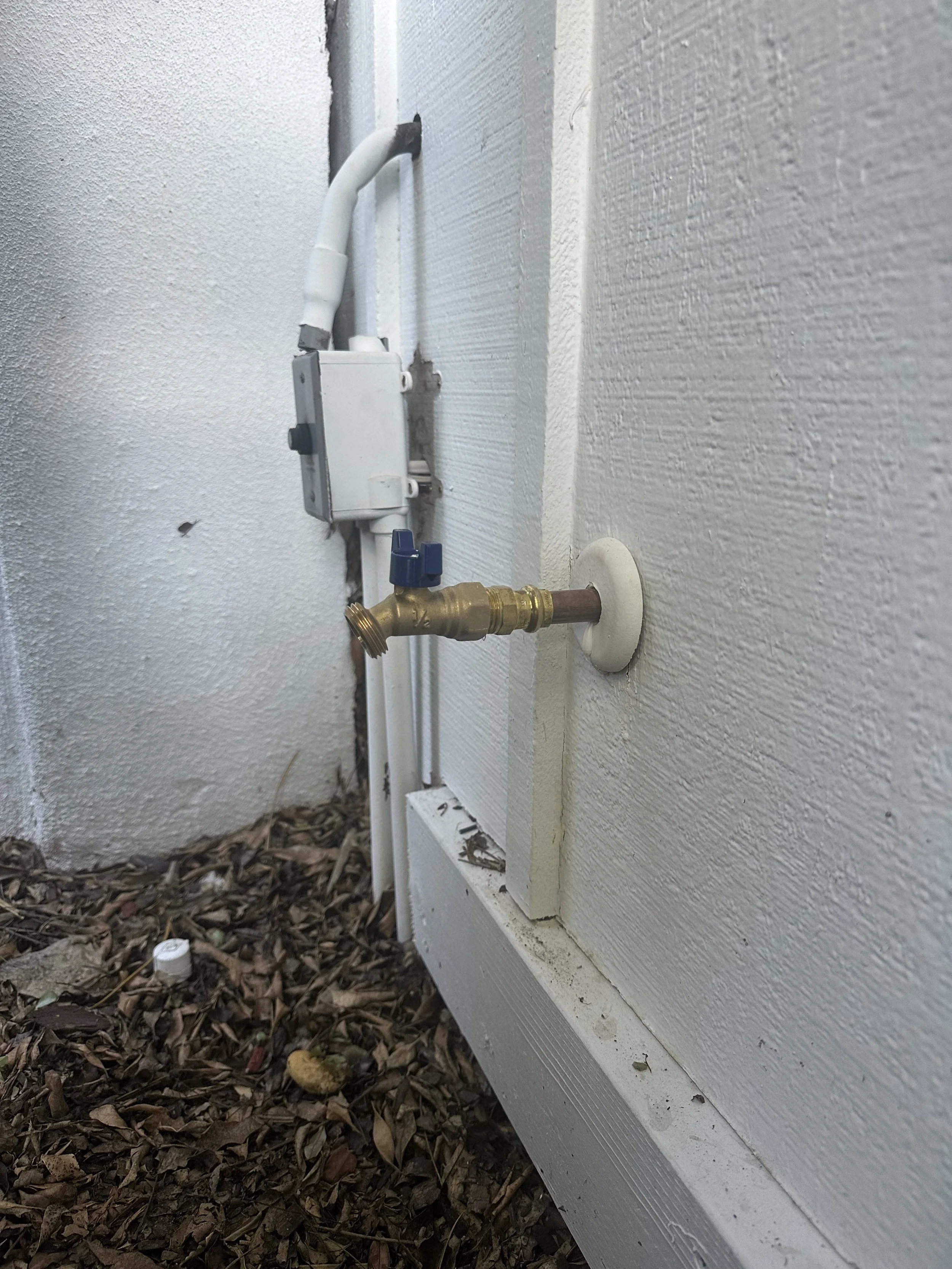 Close-up of exterior house plumbing with a brass valve attached to a pipe, and an electric water heater switch on textured white wall, on a ground covered with dry leaves.