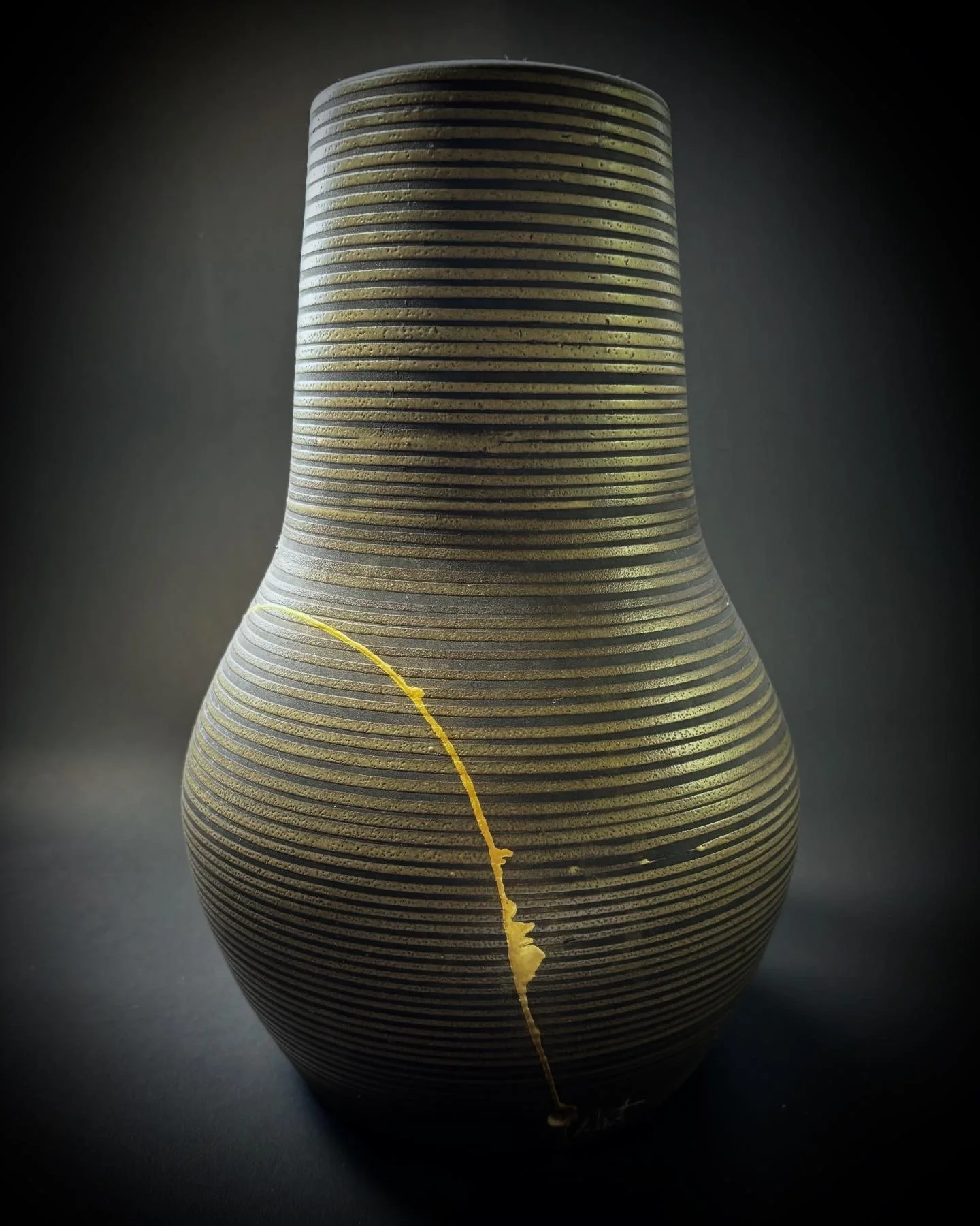 A ceramic vase with horizontal black and gold stripes, featuring a vertical crack with a gold fill on a dark background.