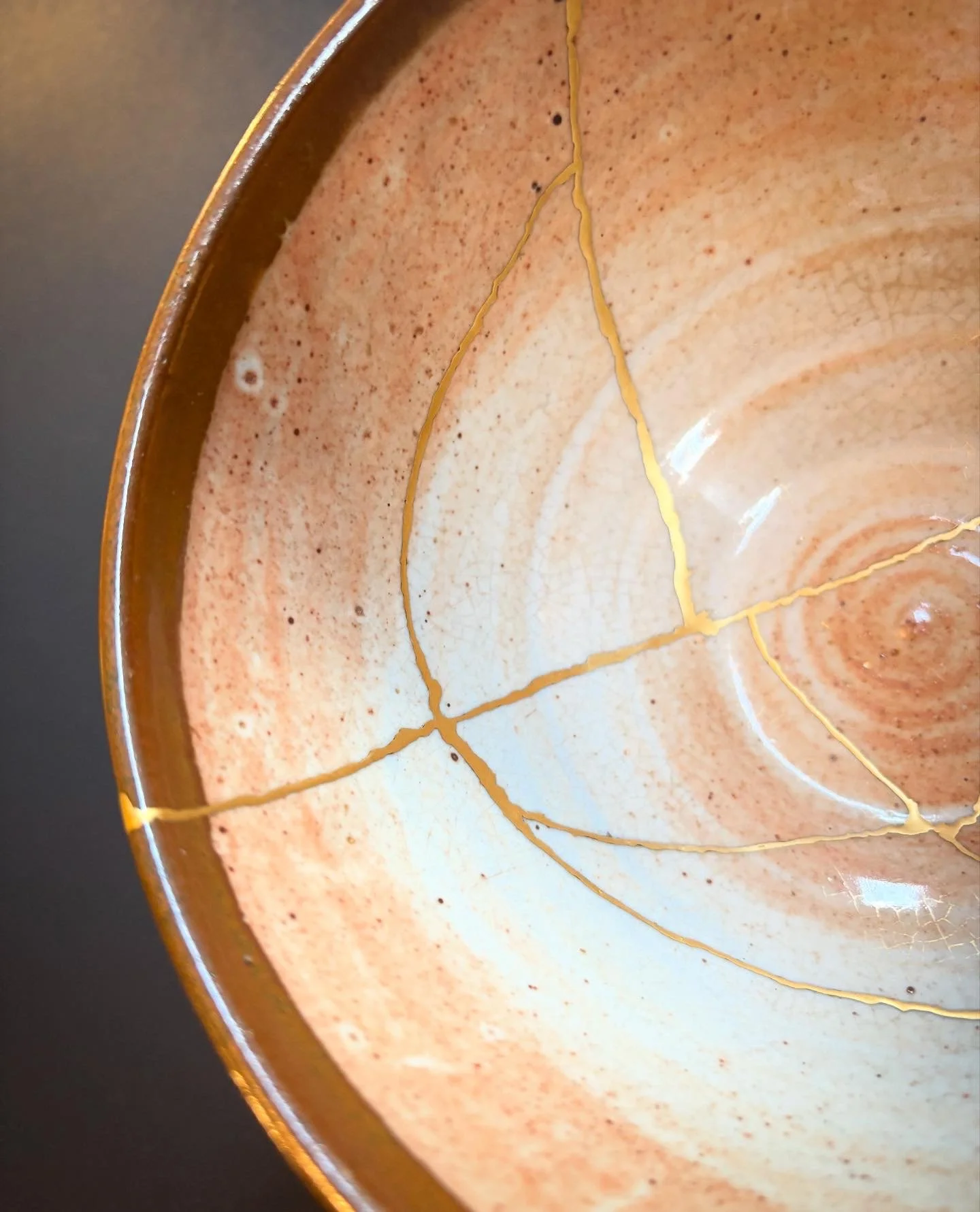 Close-up of a ceramic bowl with a tan and brown speckled glaze, showing hairline cracks filled with gold colored resin.