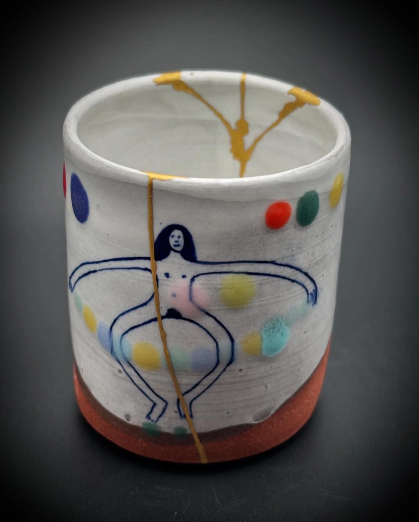 Ceramic cup decorated with colorful dots and a drawing of a woman with outstretched arms, with yellow, blue, red, and green accents, placed on a dark surface.