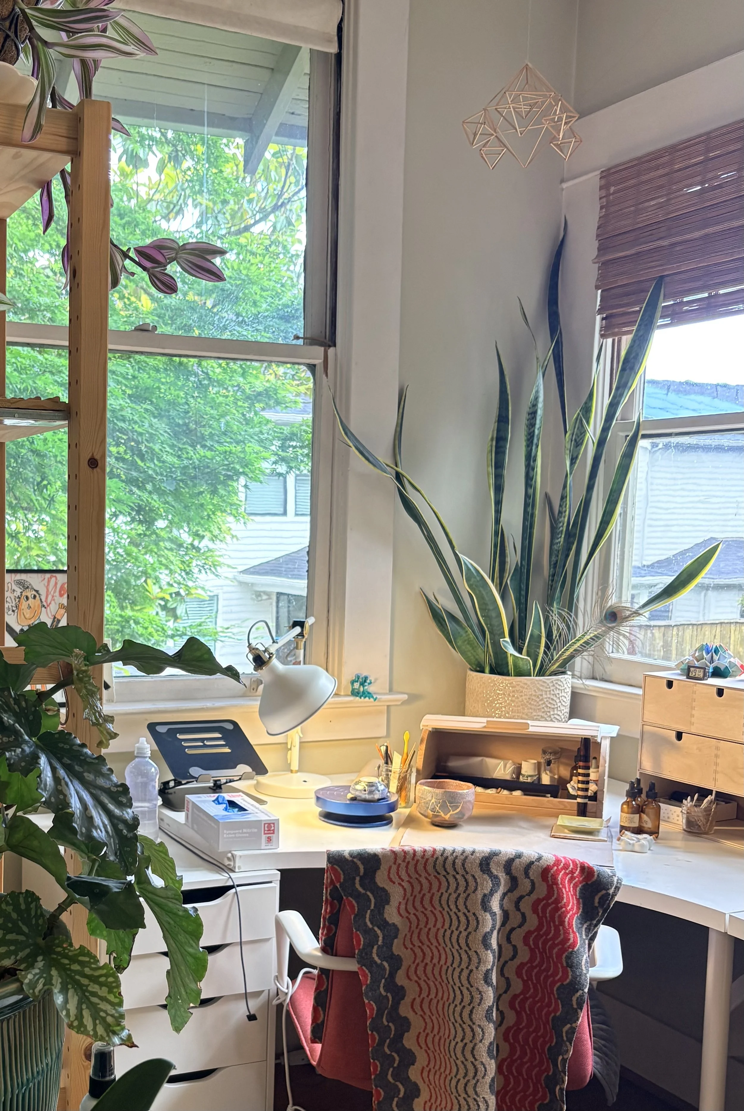 A cozy home Kintsugi art studio setup with a desk, chair, a large potted plant, a window with greenery outside, Kintsugi materials and various art and craft supplies and decorations.