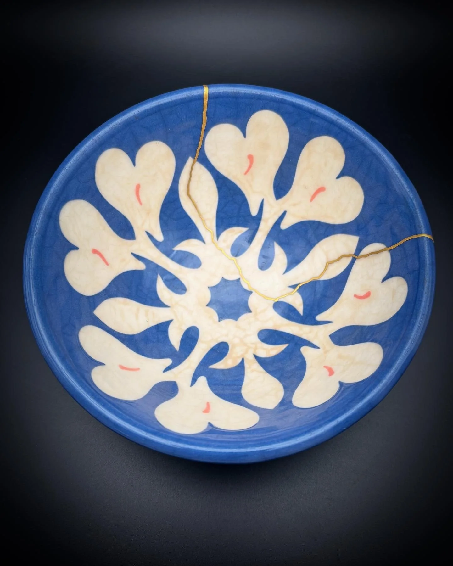 Blue ceramic plate with white floral or leaf-like pattern and tiny red accents, cracked with two visible fractures.
