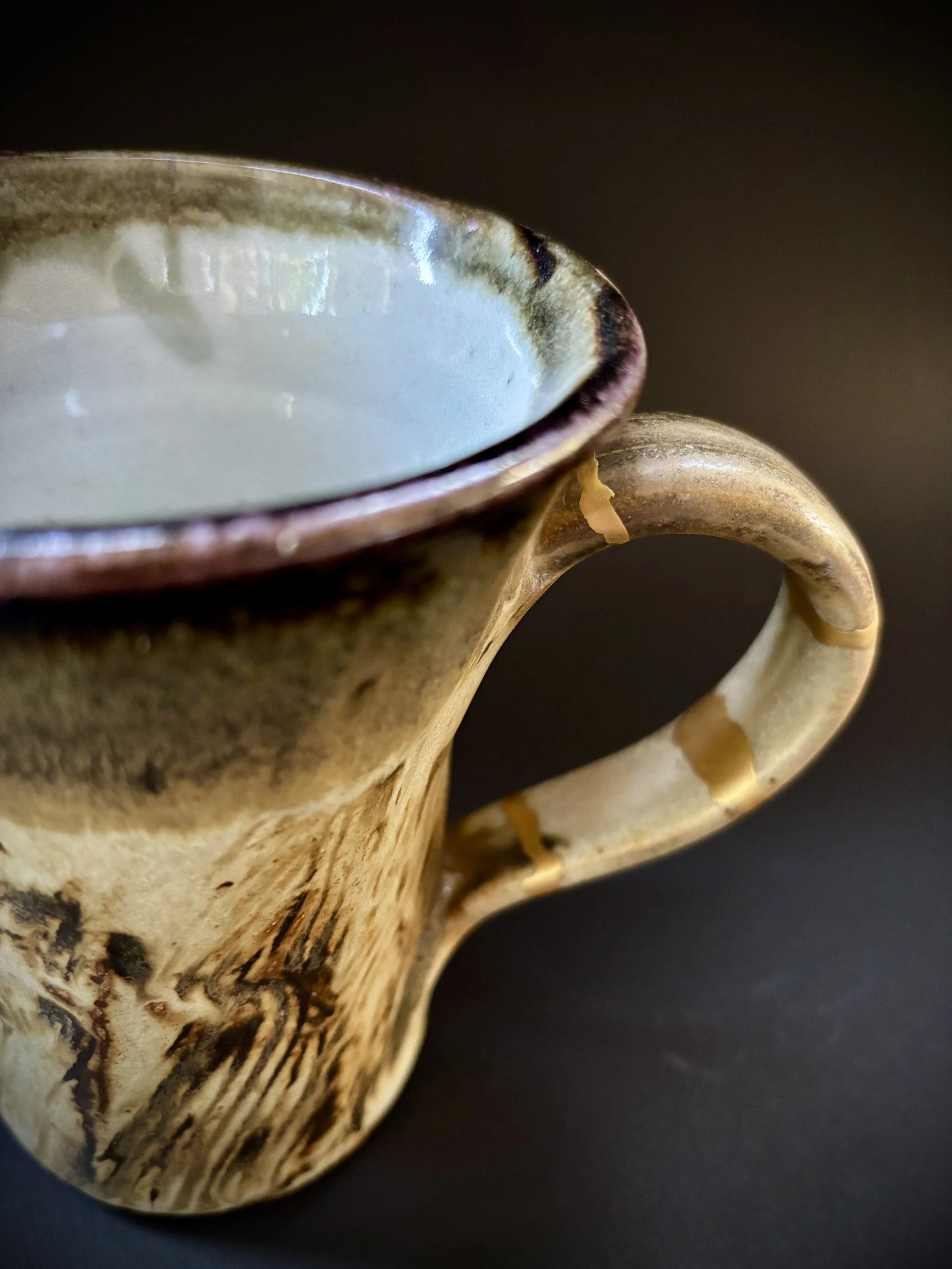 Close-up of a ceramic mug with a beige and brown glazed finish, mounted with a smooth curved handle, atop a dark surface.