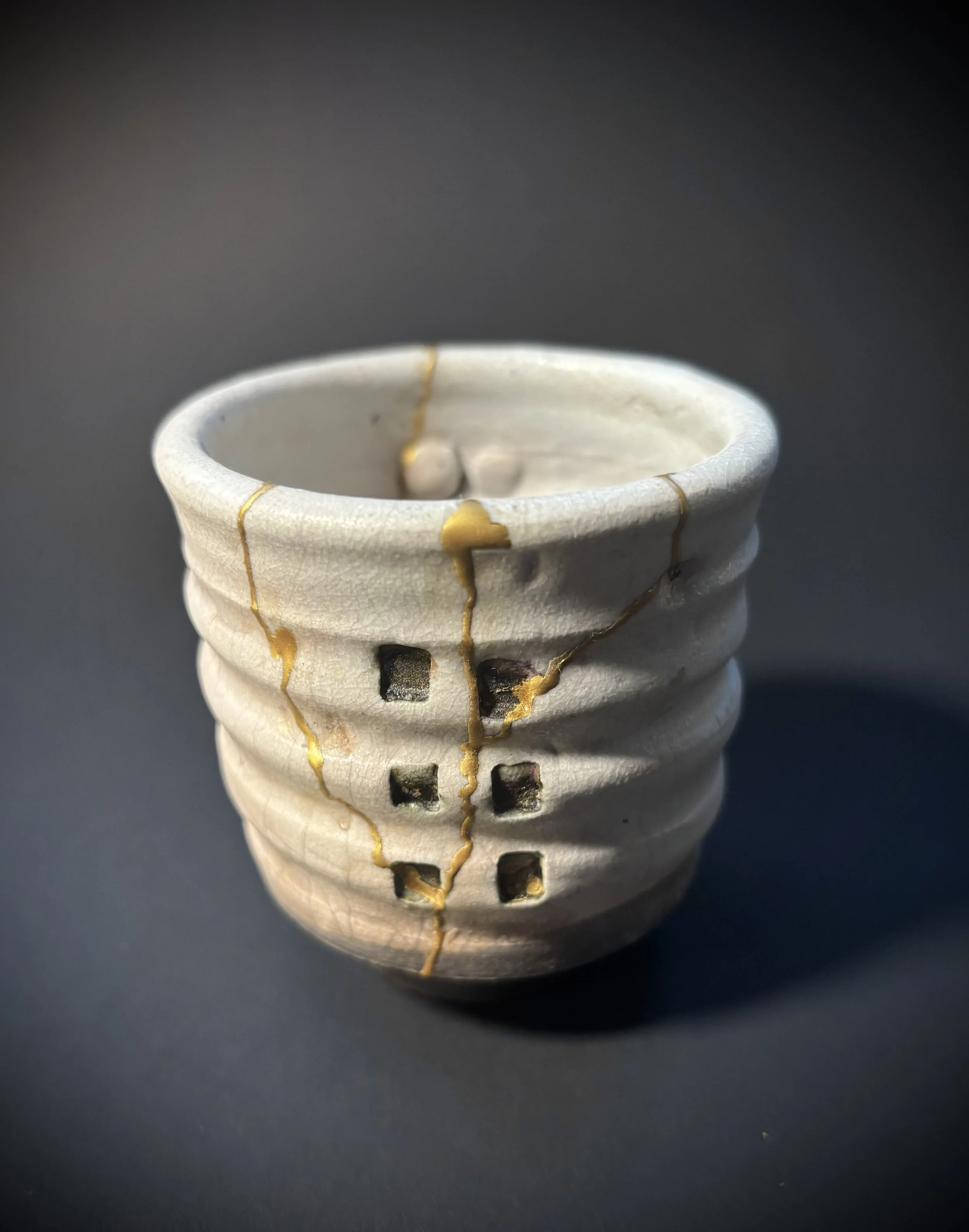 Close-up of a small, handcrafted ceramic cup with gold and black square accents, placed on a dark surface.