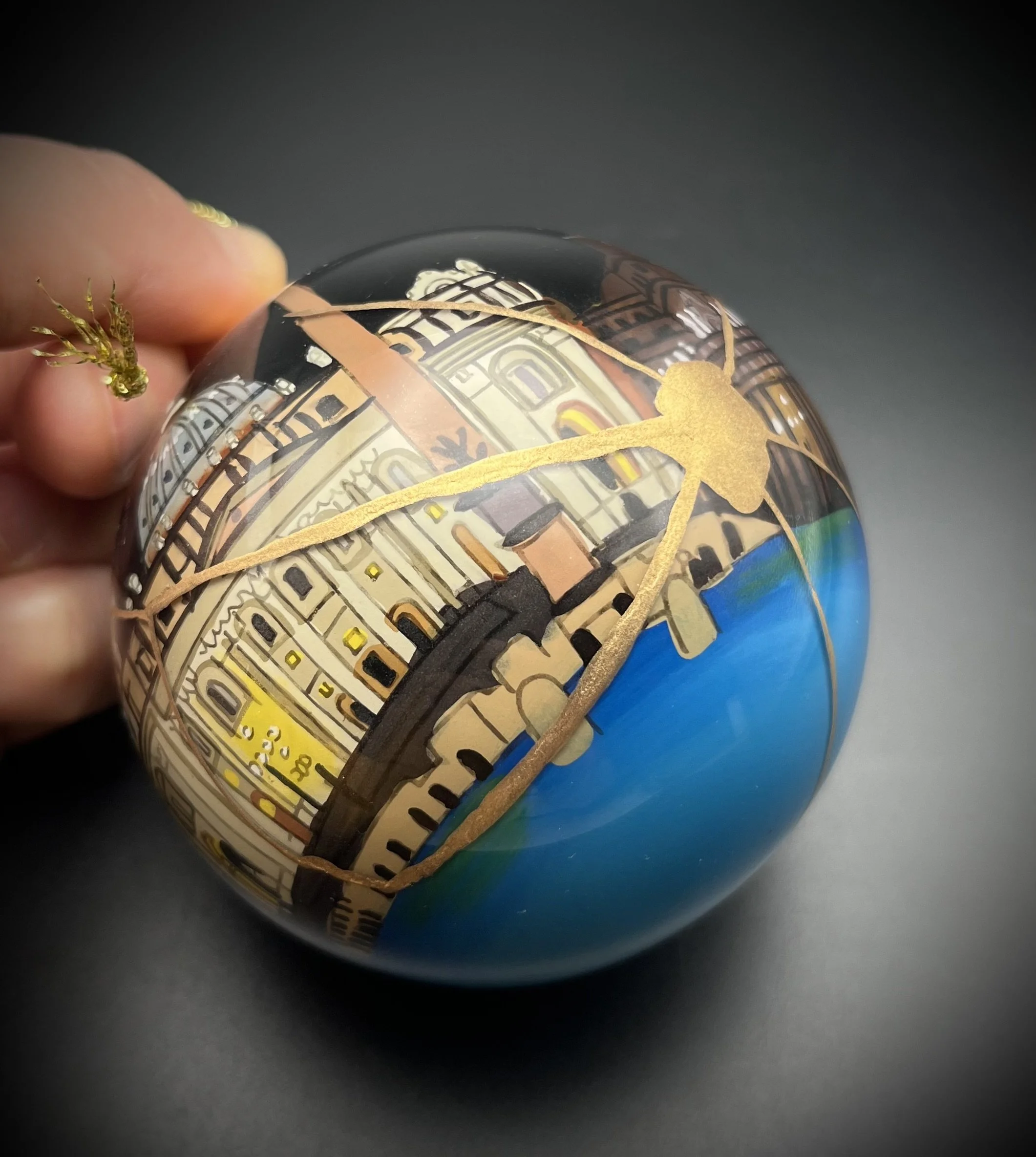 Close-up of a decorative glass ornament featuring colorful illustrations and gold accents, held by a person's fingers against a dark background.