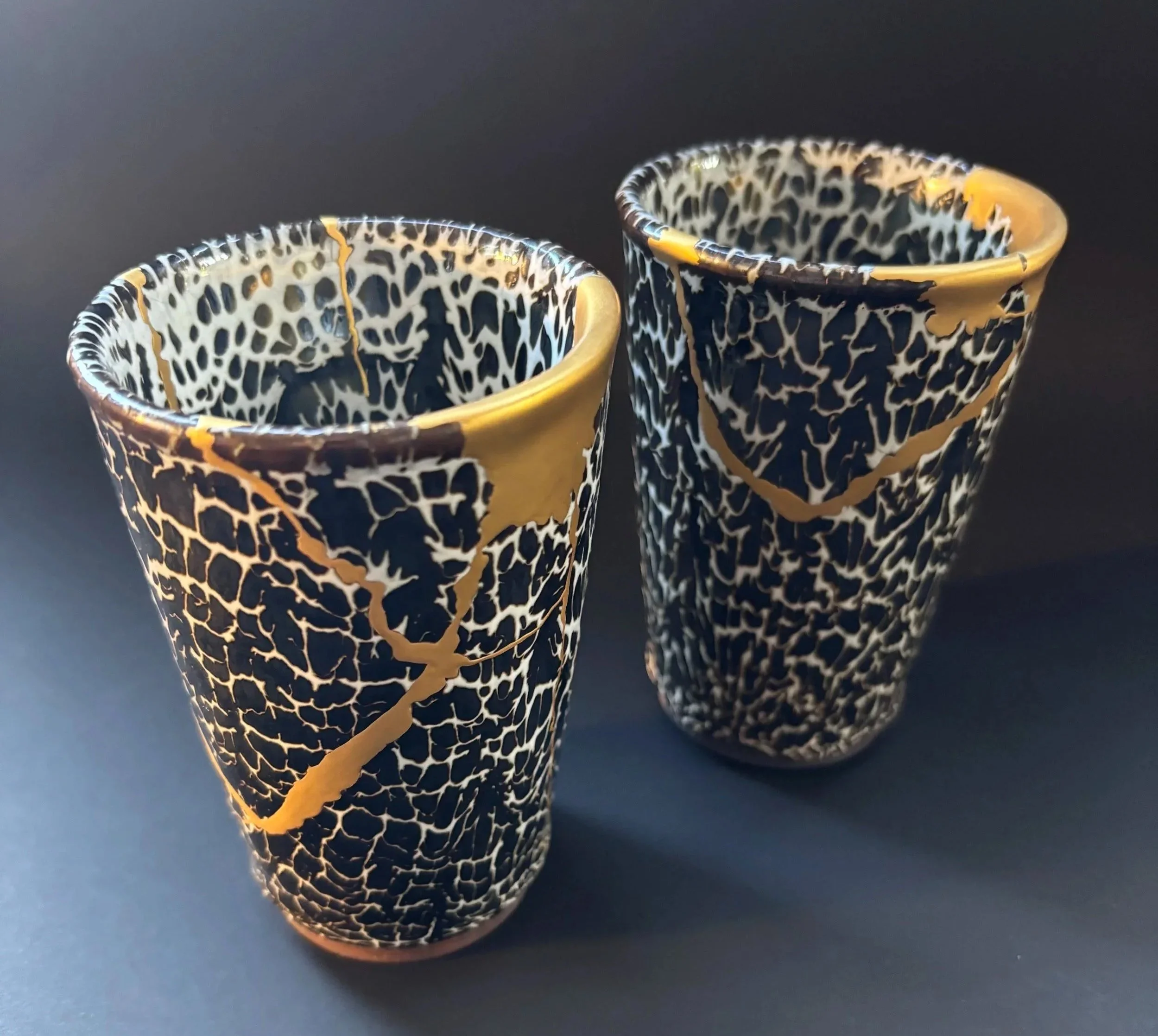 Two black ceramic cups with intricate white and gold crackle patterns on a dark surface.