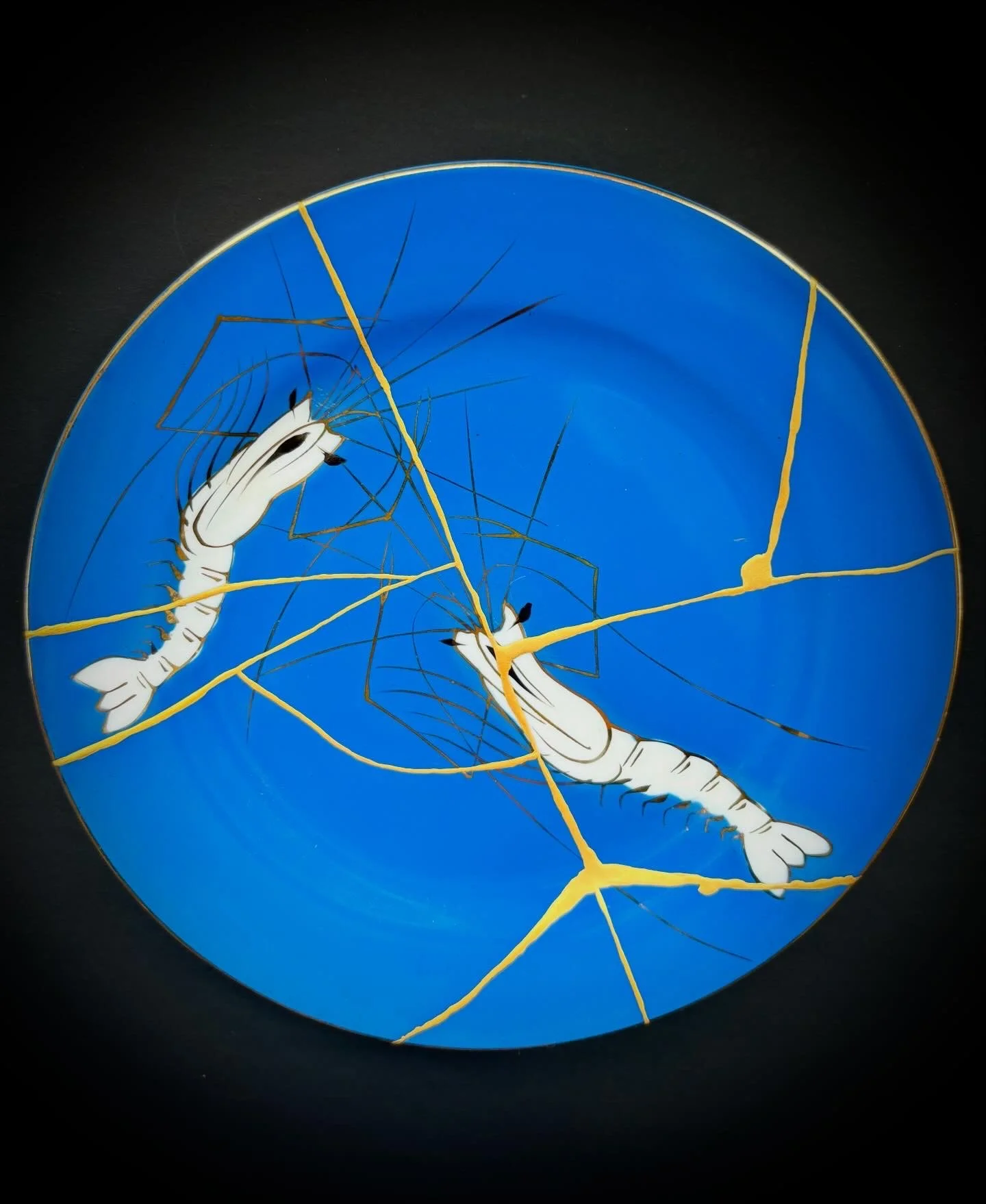 A circular decorative plate featuring a blue background with a hand-painted illustration of a white shrimp with black eyes and long antennae. The plate has cracks with irregular gold lines running across it, creating a crackled effect.