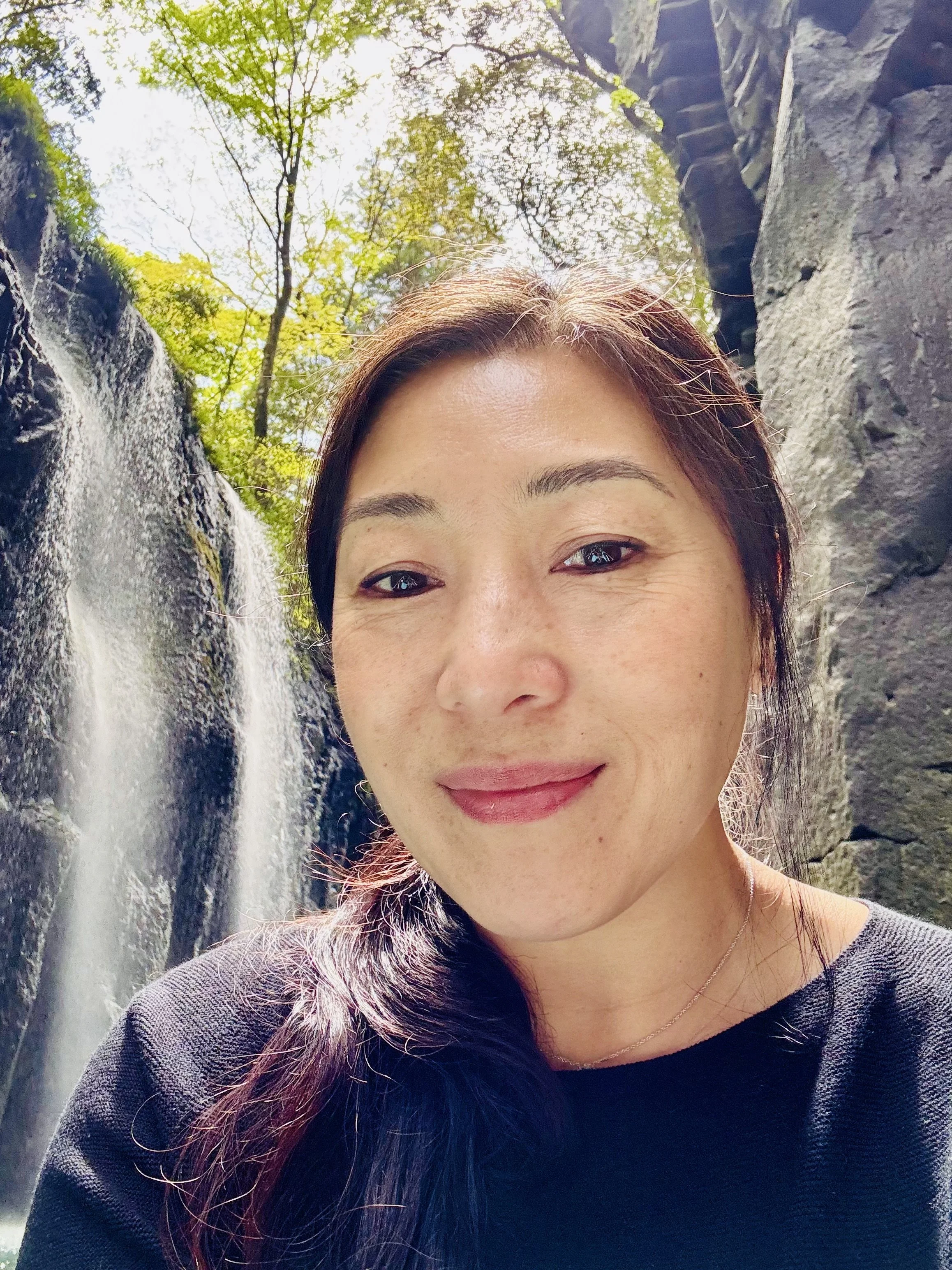 A woman with long dark hair and a slight smile taking a selfie outdoors near a waterfall with lush green trees in the background.