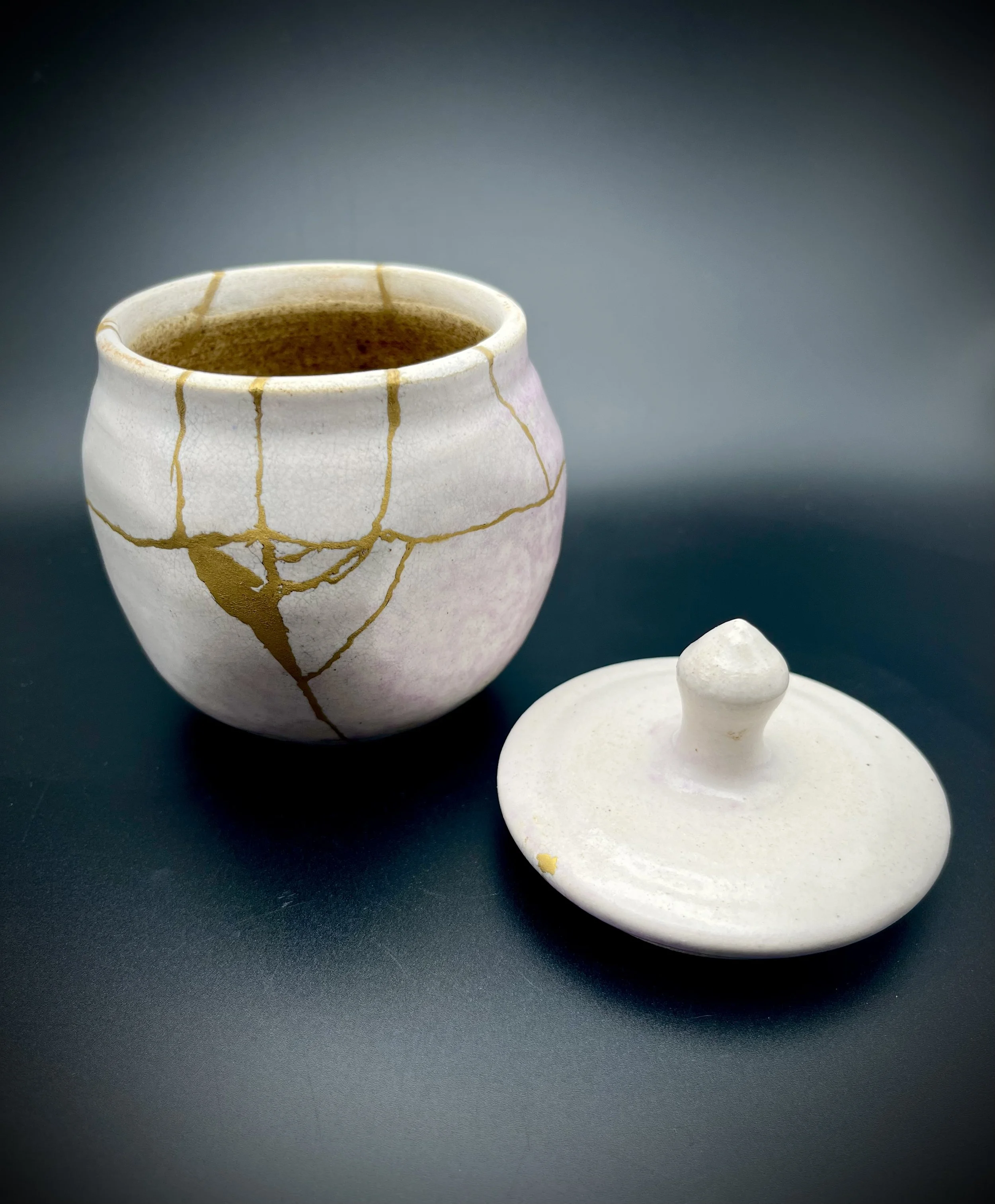 Close-up of a white ceramic tea or sugar container with a crackled glaze and gold lines, along with a matching lid on a dark surface.