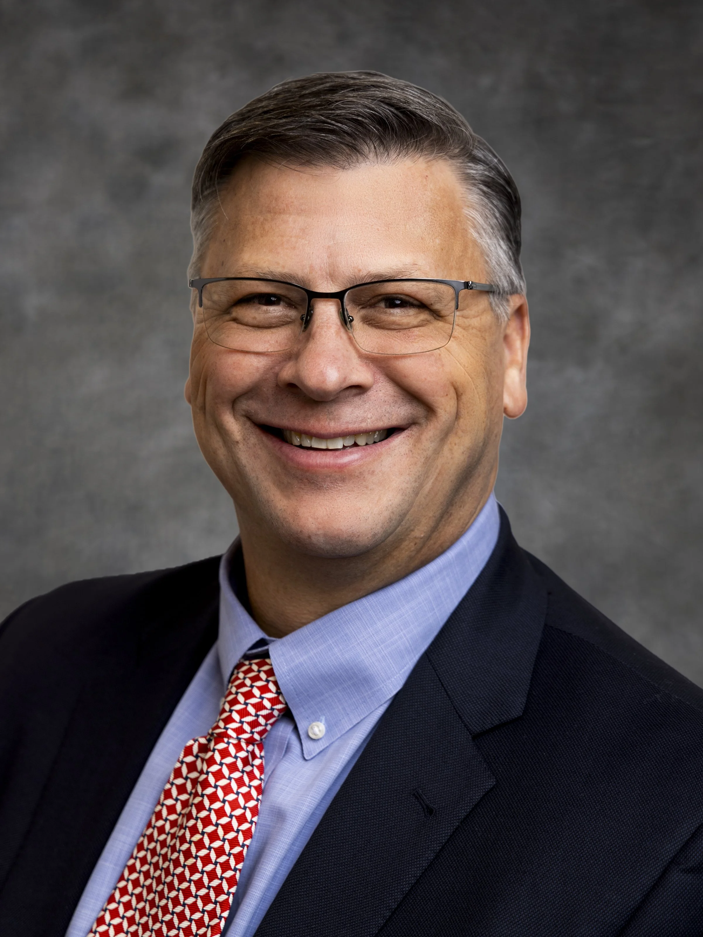 Smiling middle-aged man with glasses wearing a suit, tie, and dress shirt, professional portrait, gray background.