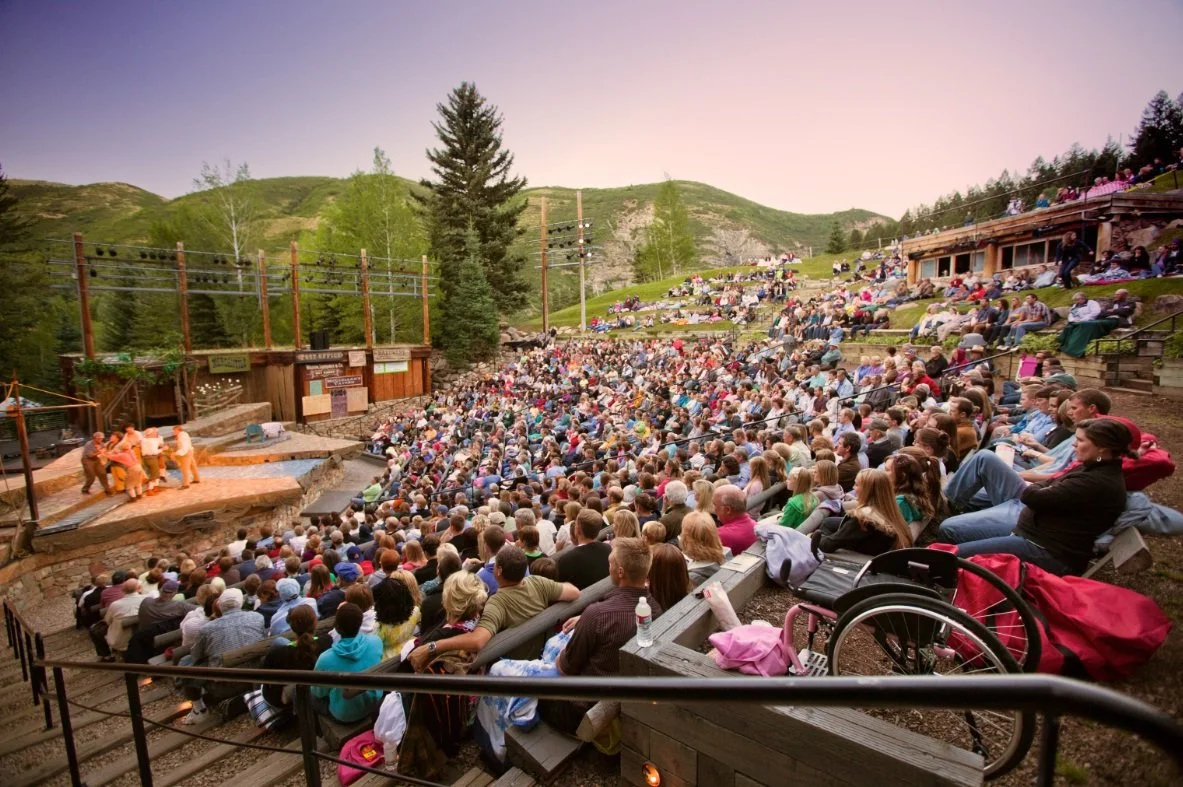 An outdoor theater with a large audience watching a performance at sunset in a mountainous area surrounded by trees.