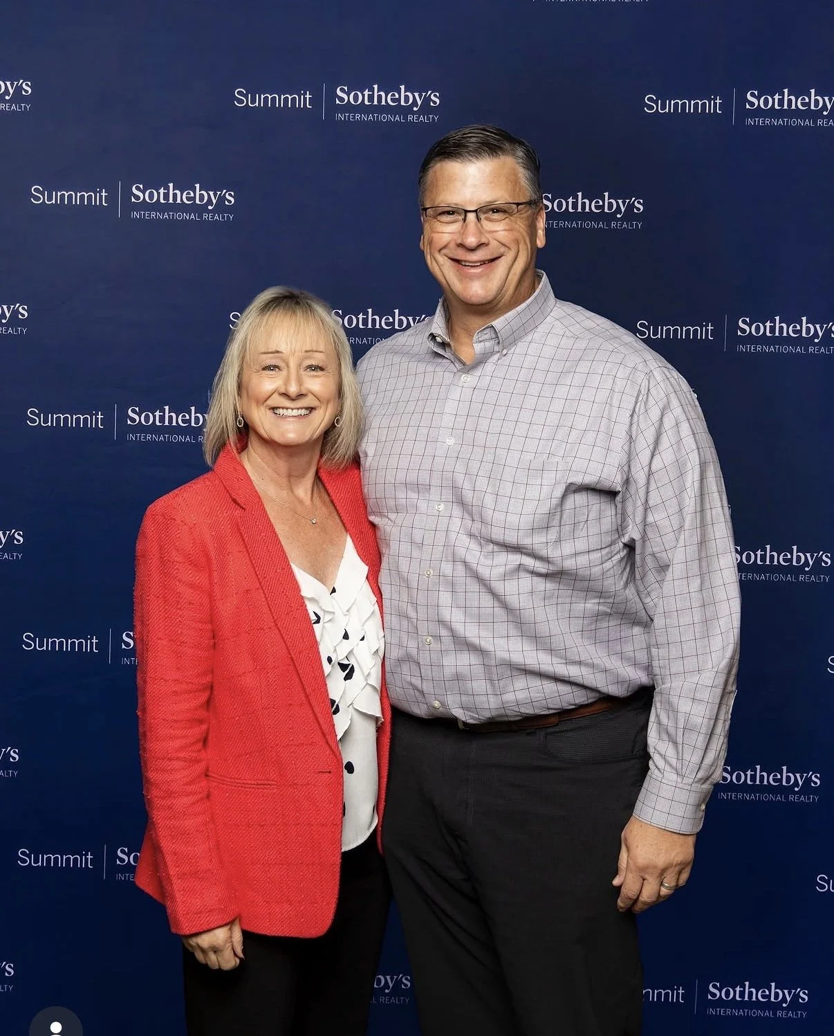 A smiling woman in a red blazer and a smiling man in a gray checkered shirt standing together at a Sotheby's International Realty event with a blue backdrop featuring the Sotheby's and Summit logos.