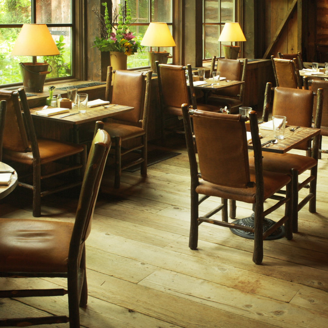 A rustic restaurant interior with wooden tables and leather chairs, decorated with lamps and a flower arrangement, near large windows showing greenery outside.