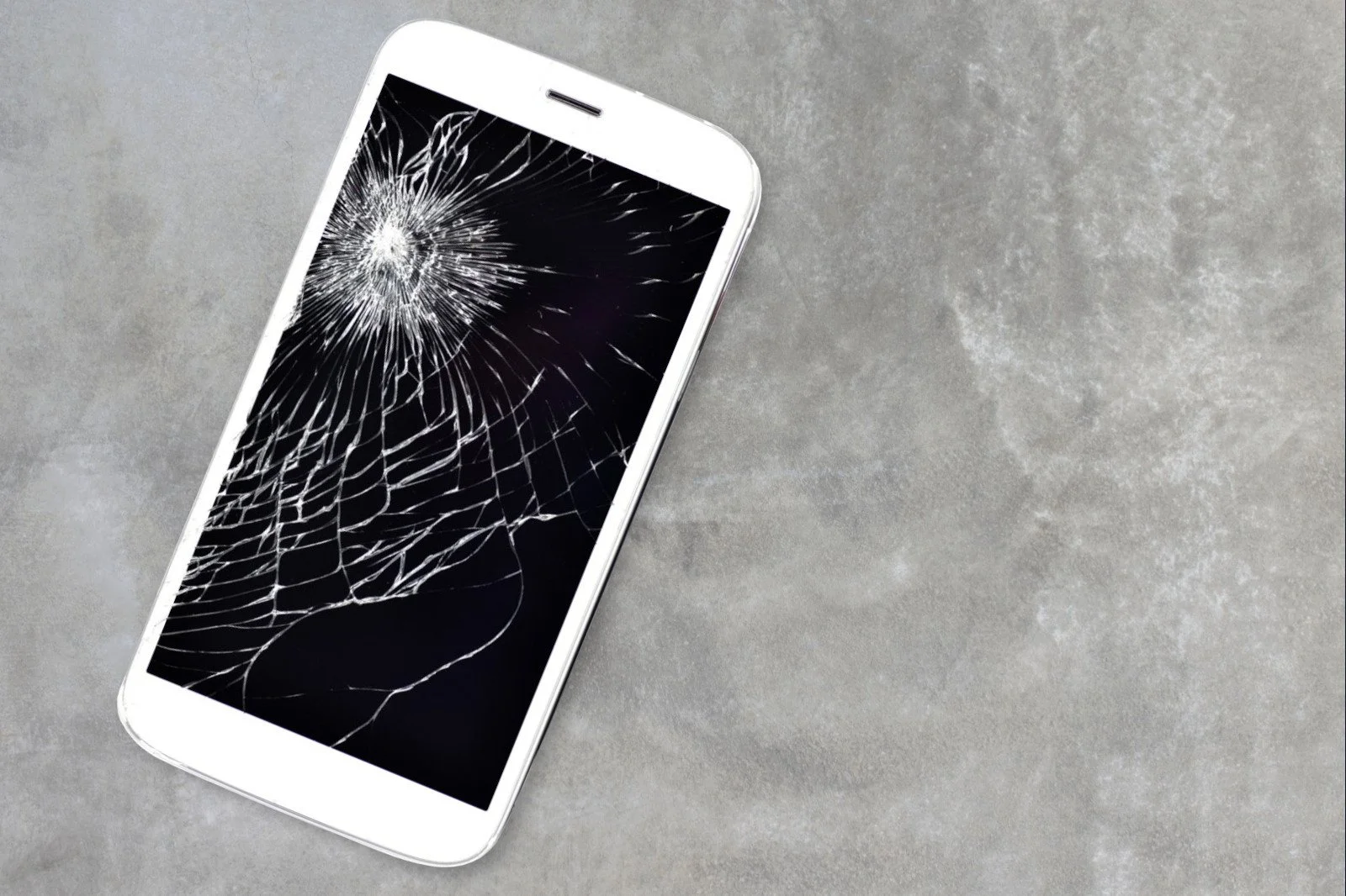 A smartphone with a shattered screen lying on a gray surface.