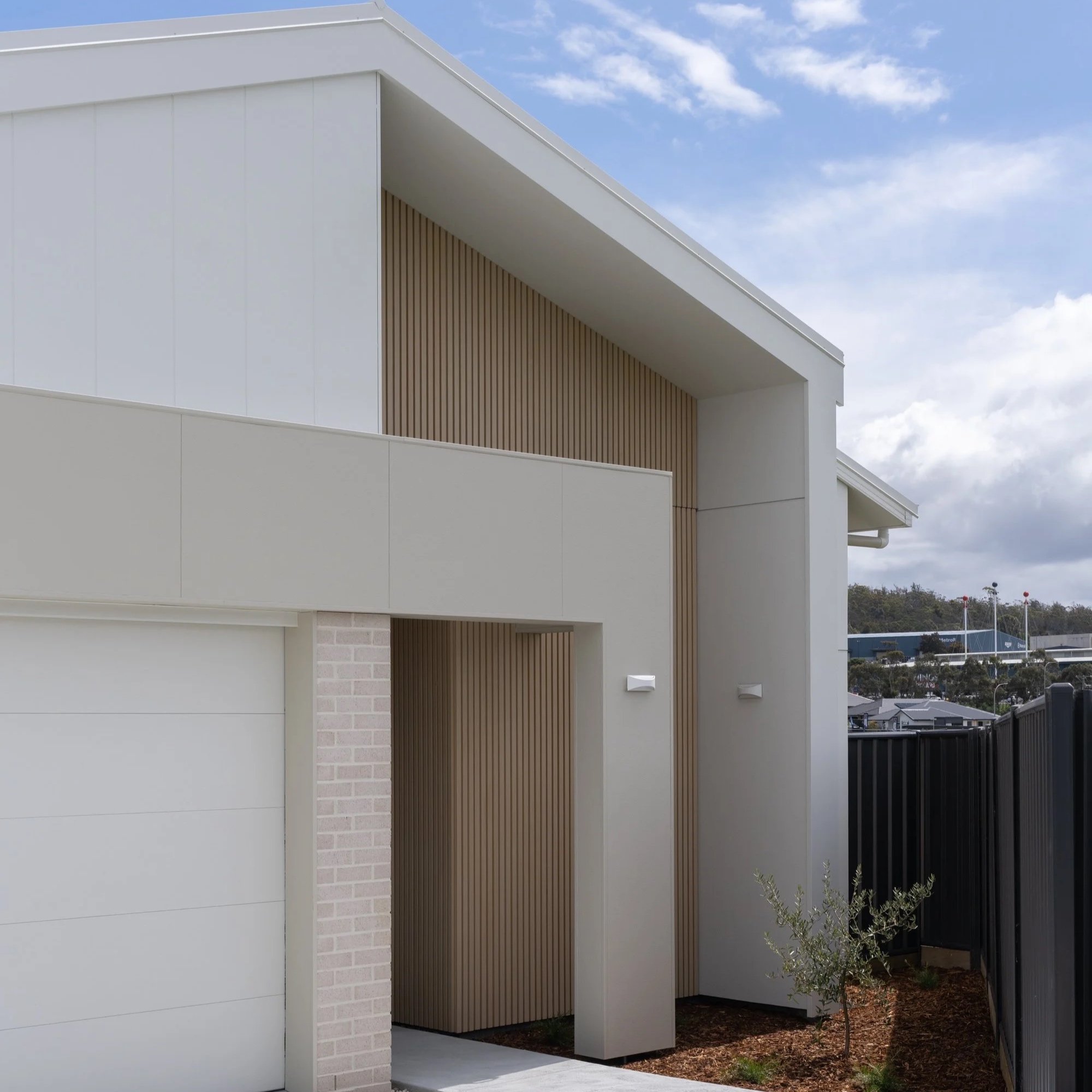 Modern white building with a slanted roof, corner details, and wooden accent wall, under a partly cloudy sky. A small landscaped area with trees and a fence is visible in the background.