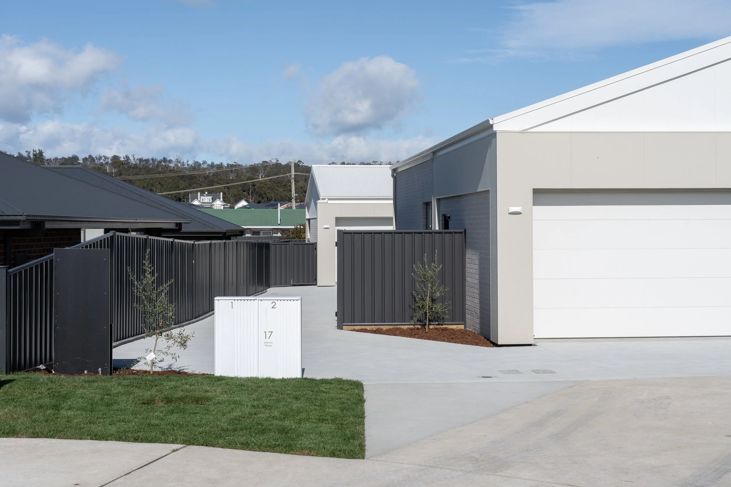 Modern residential town-houses with black fences, a white mailbox, and a small tree on a concrete driveway. Clear blue sky with scattered clouds in the background.